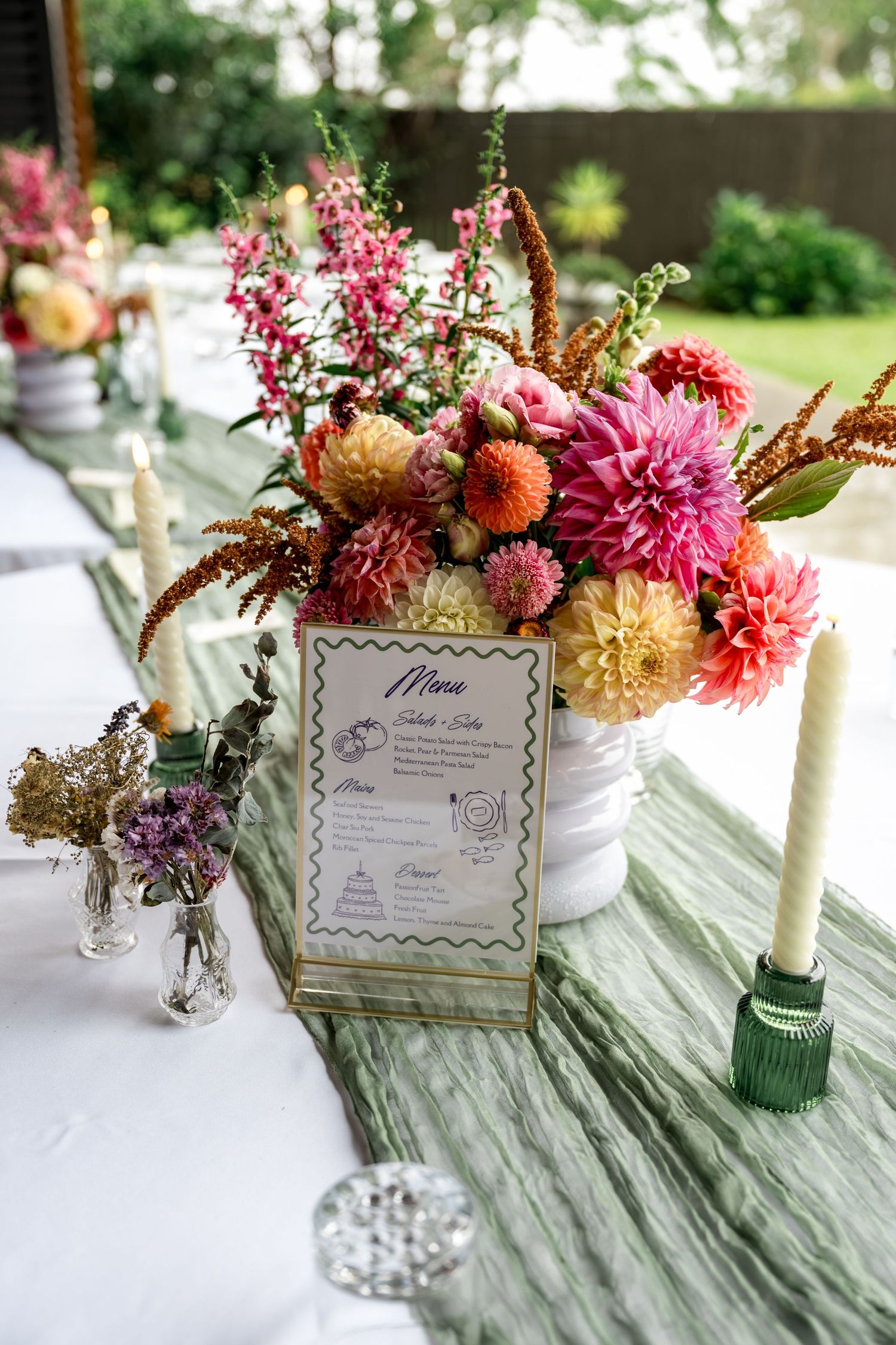 Wedding reception table with colorful dahlia centerpiece, menu card, taper candles, and green runner.