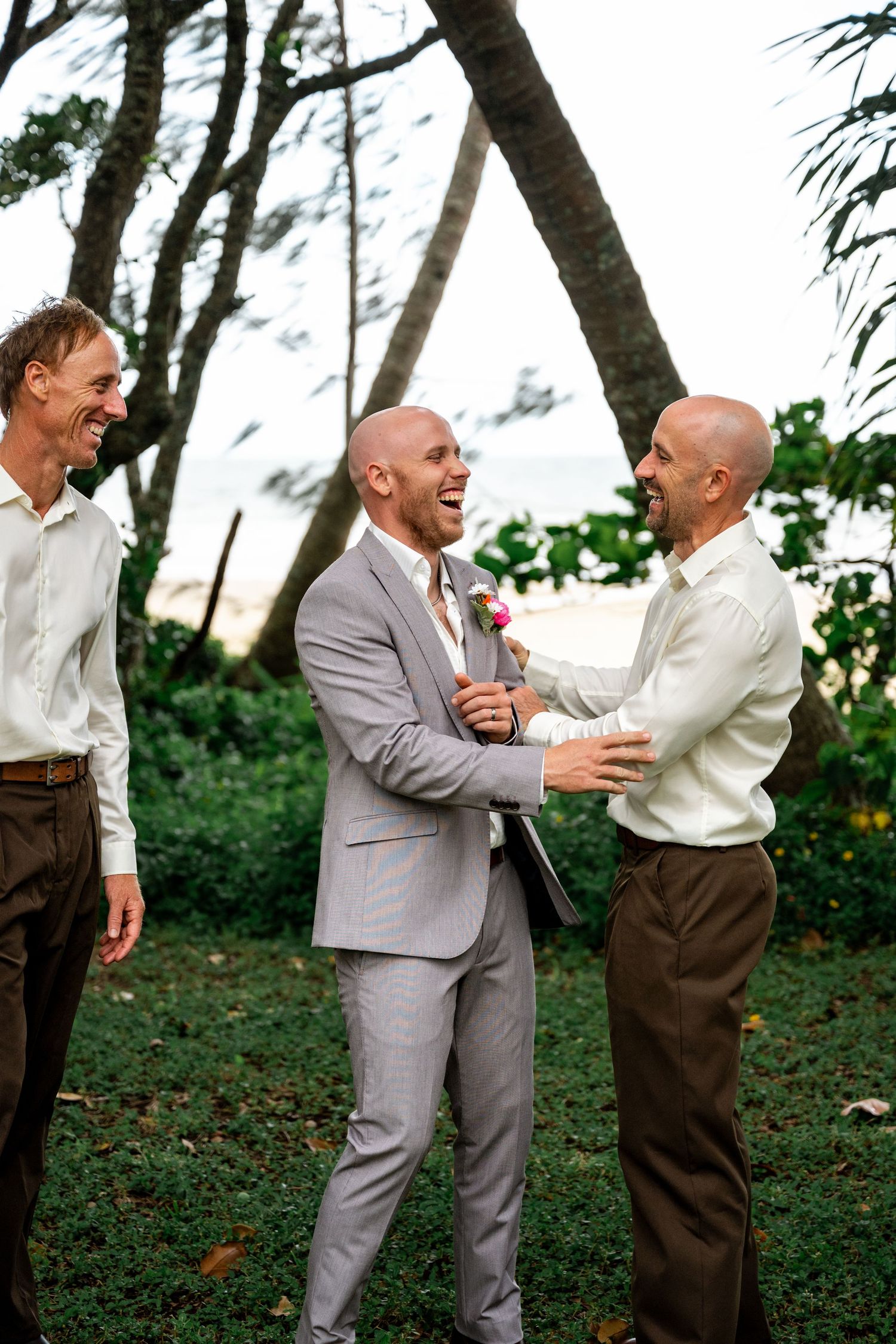 Groom in grey suit shares joyful laugh with groomsmen in white shirts during outdoor tropical wedding celebration.