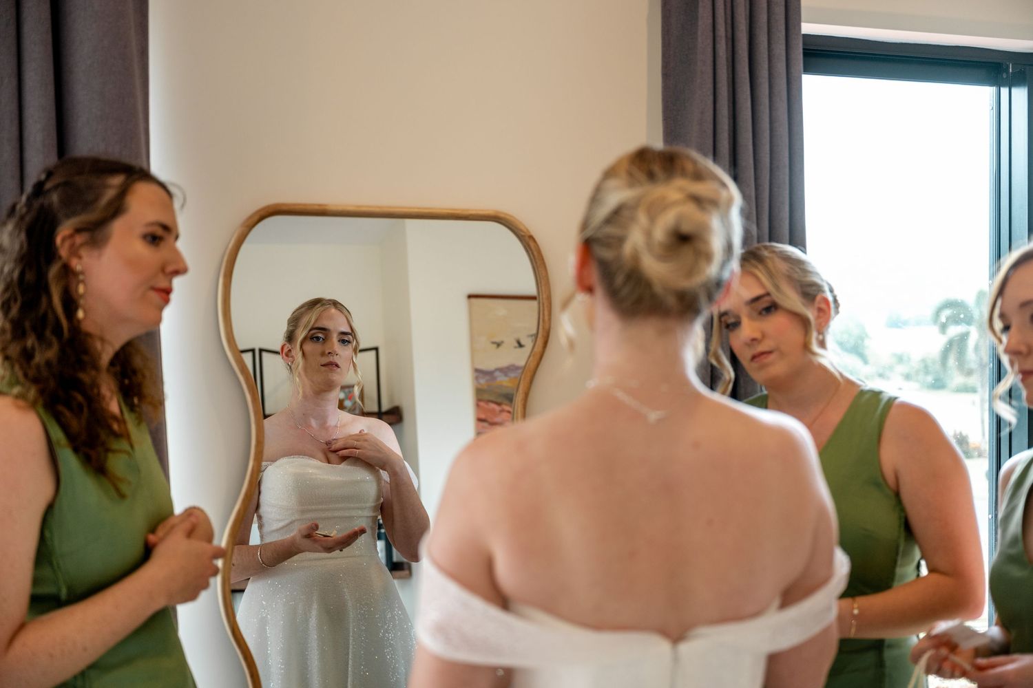 Bride views her wedding dress in mirror surrounded by bridesmaids in sage green gowns.