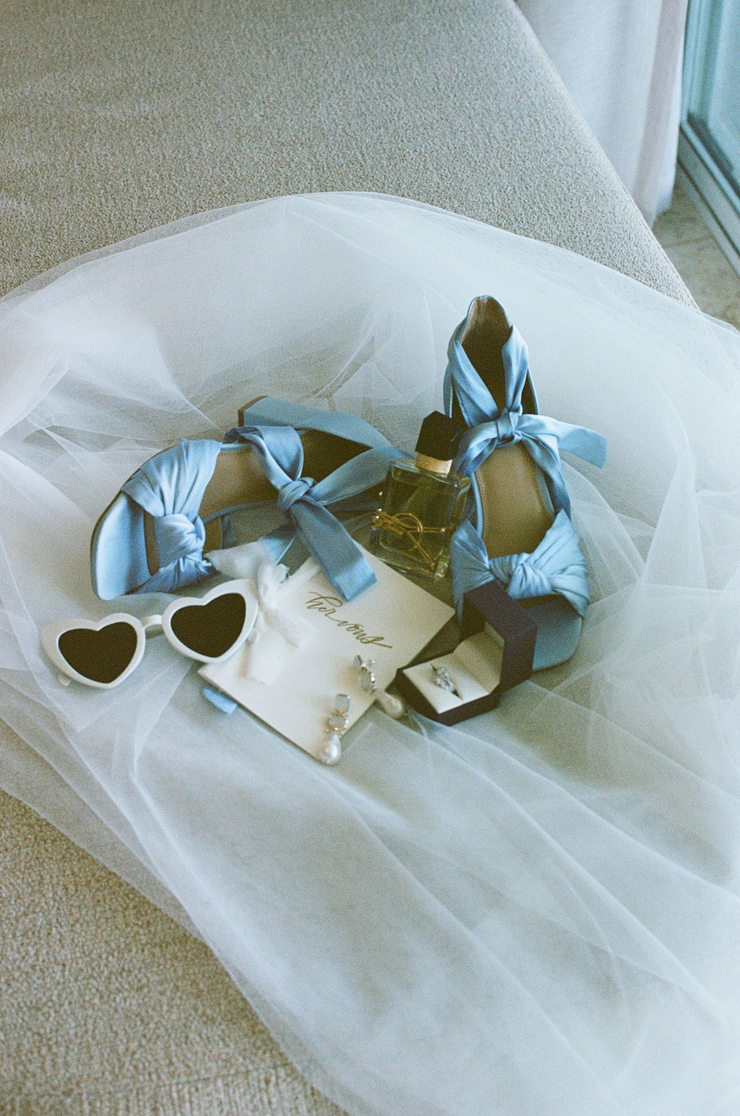 Light blue satin bridal heels with bow ties beside heart sunglasses and perfume on white tulle.
