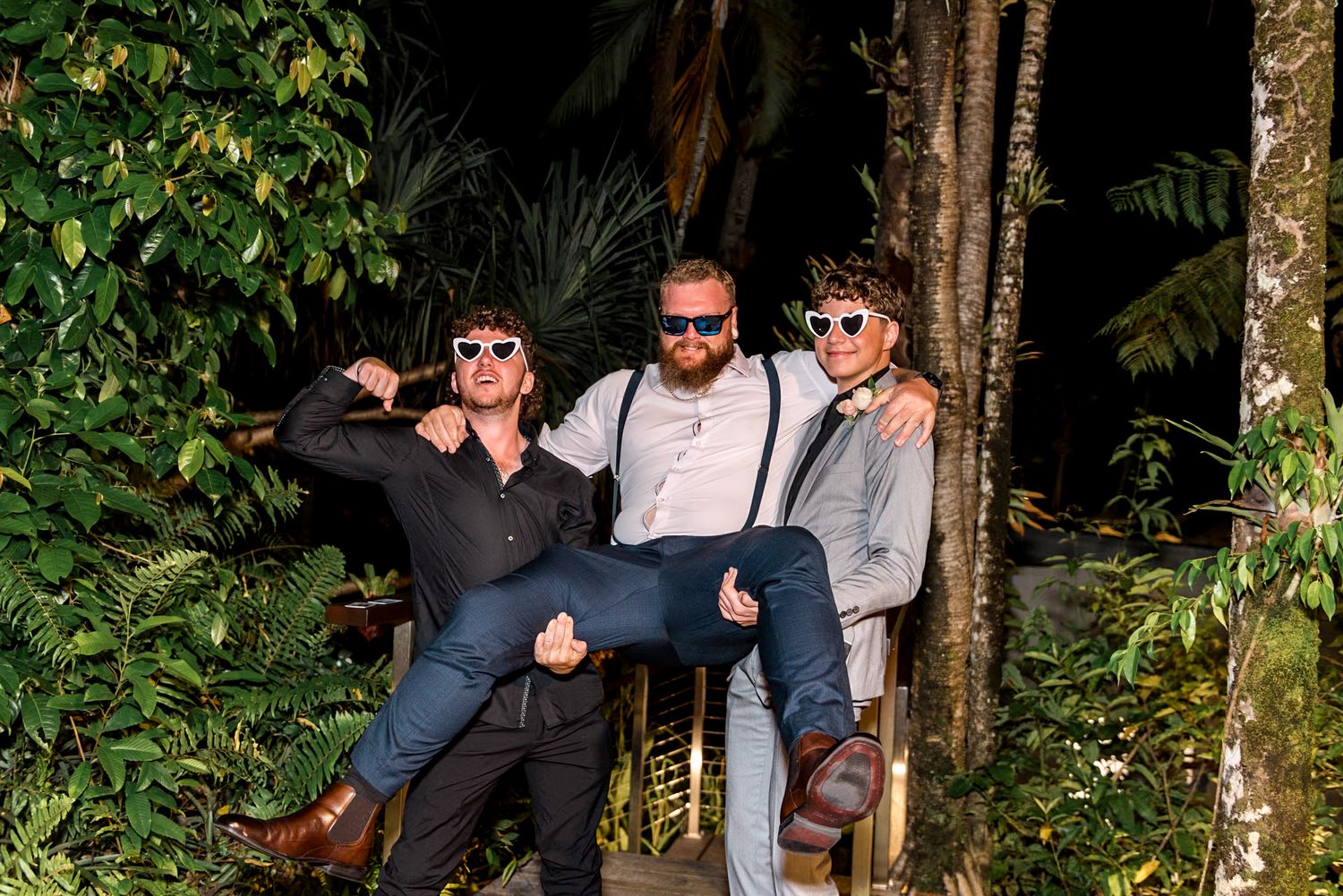 Three men in formal attire posing playfully among tropical green foliage wearing sunglasses.