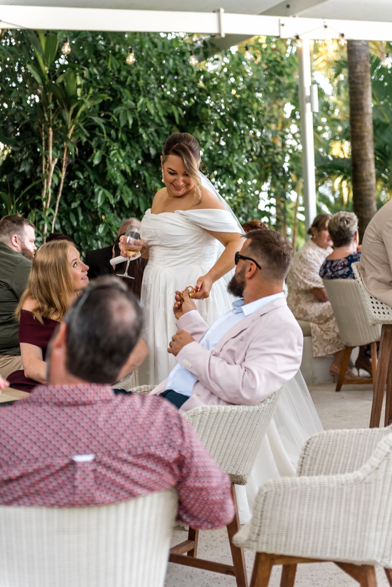 Smiling bride in off-shoulder gown interacts with seated groom at outdoor wedding reception.