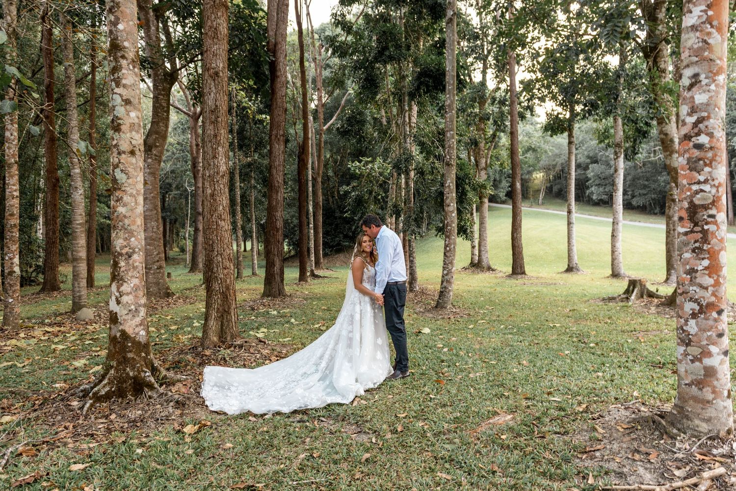 Bride and groom embrace intimately among tall trees on a lush green lawn in a forest setting.