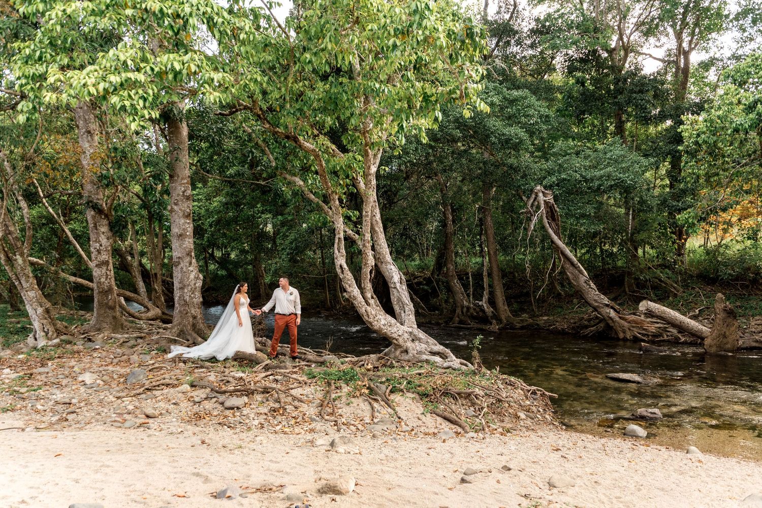 Bride and groom hold hands on a sandy riverbank surrounded by lush forest trees.