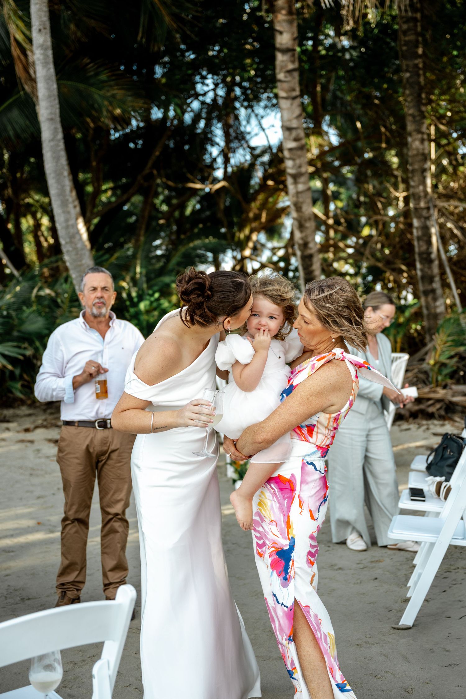 Bride in white gown kissing a young child held by a woman in floral dress at beach wedding at Four Mile Beach, Port Douglas.