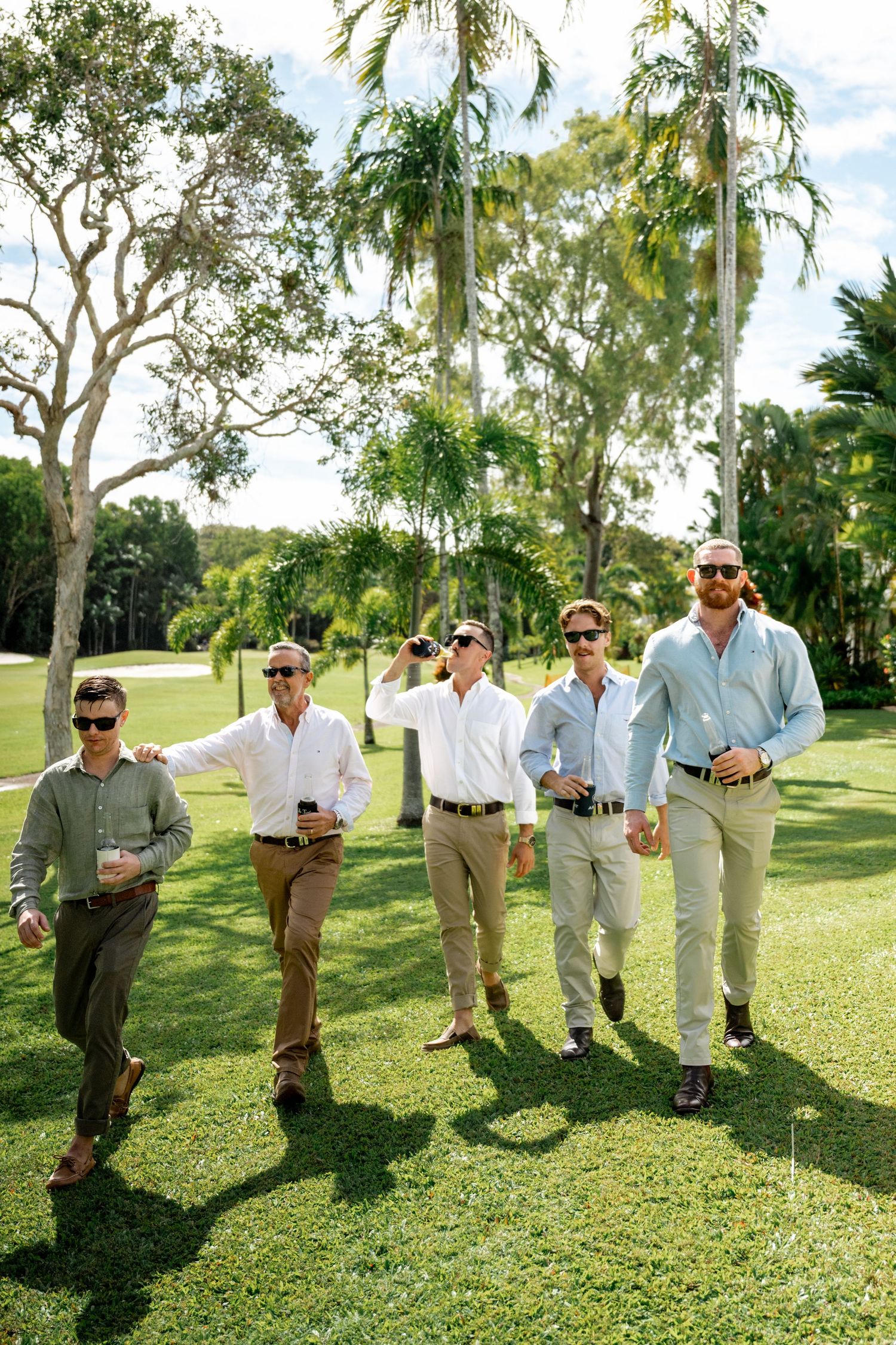 Six groomsmen in sunglasses and casual shirts walking confidently across a tropical golf course lawn.