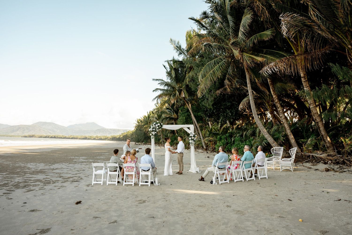 Wide shot of small intimate beach wedding ceremony with guests seated facing couple at white floral arch at Four Mile Beach, Port Douglas.