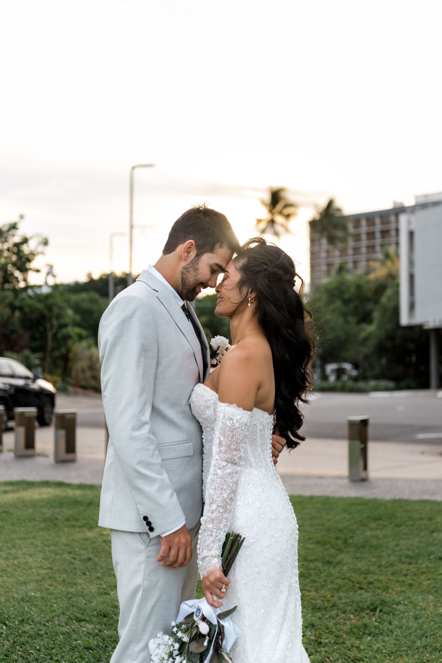 Bride and groom touch foreheads intimately at sunset on a green lawn in elegant wedding attire.