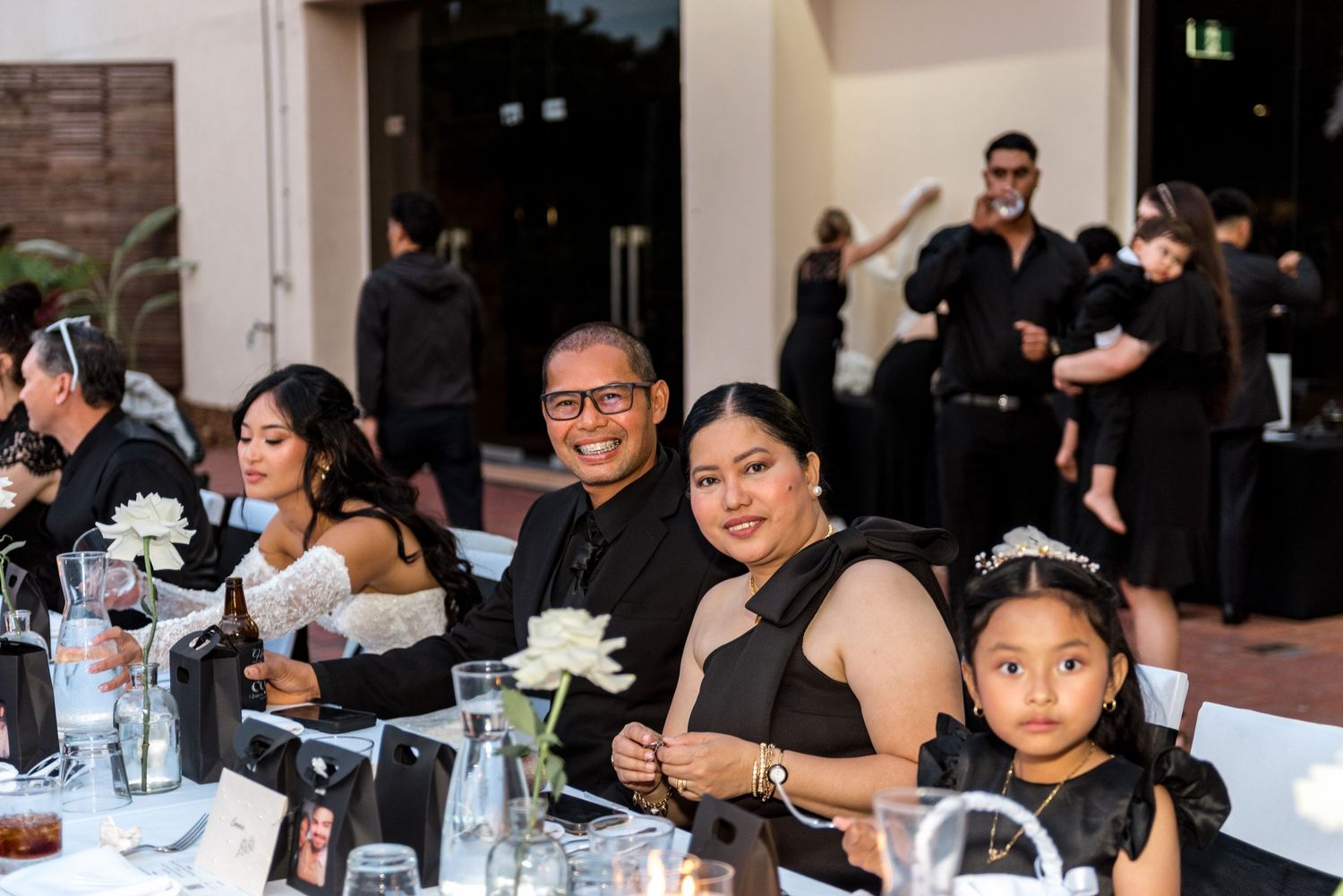 Wedding guests in black attire smile at the camera while seated at a decorated reception table with white flowers.