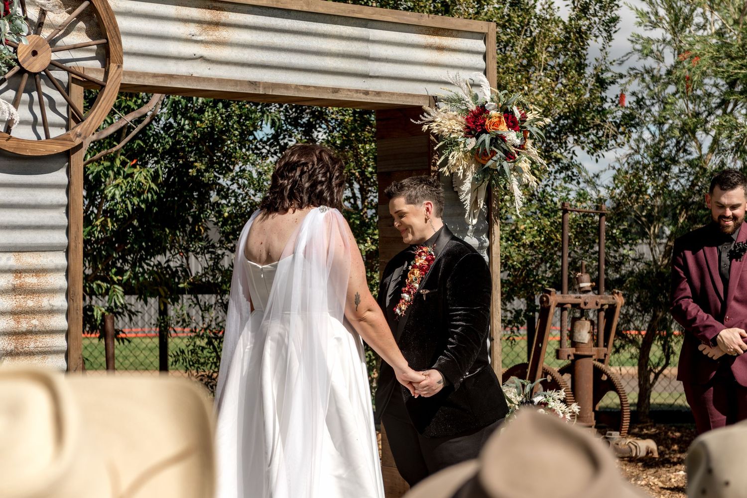 Bride and groom hold hands facing each other at rustic outdoor wedding altar with colorful floral decor.