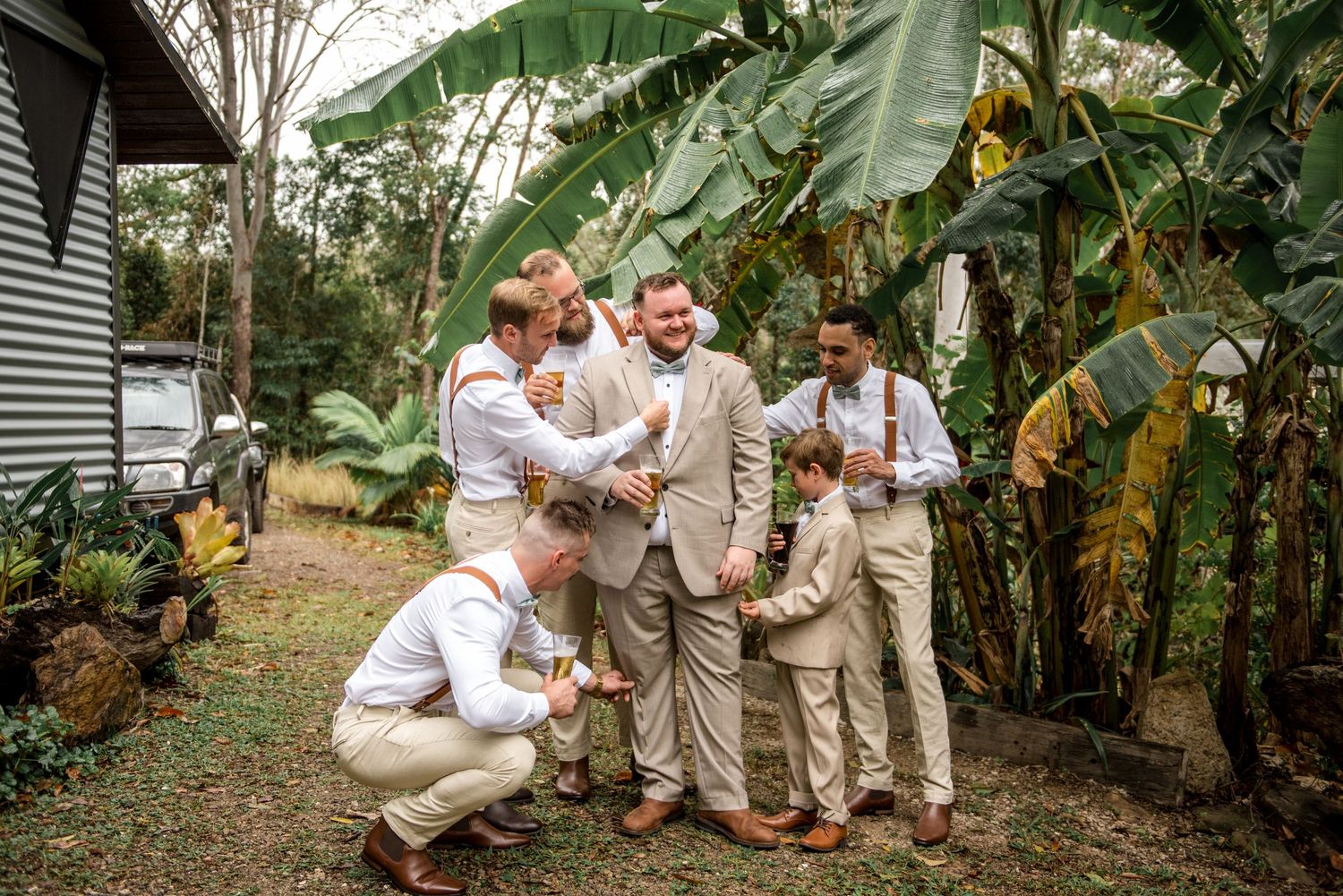 Groomsmen and ring bearer laughing together in lush tropical garden before wedding ceremony.