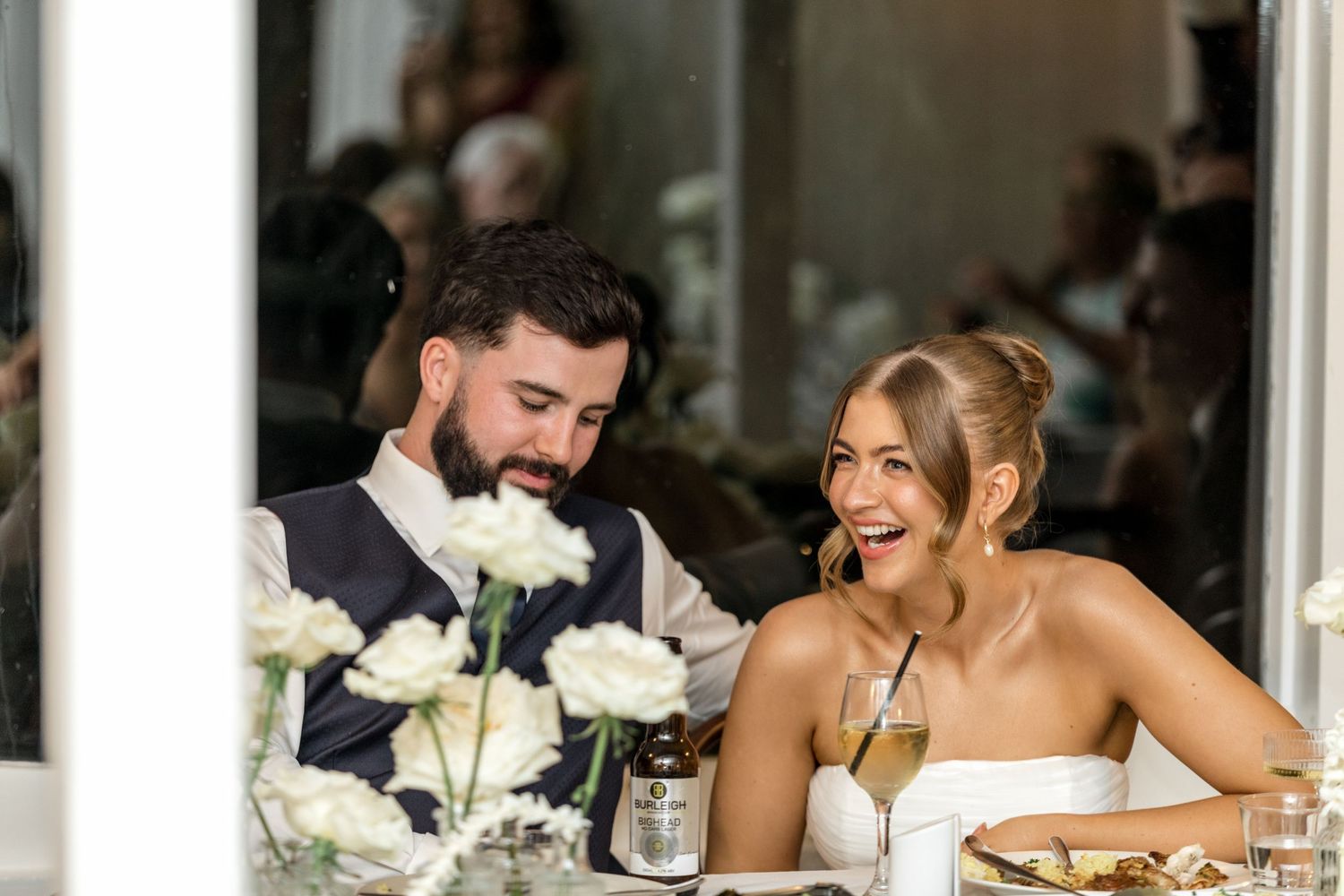 Bride and groom laughing together at wedding reception table with white floral centerpiece.