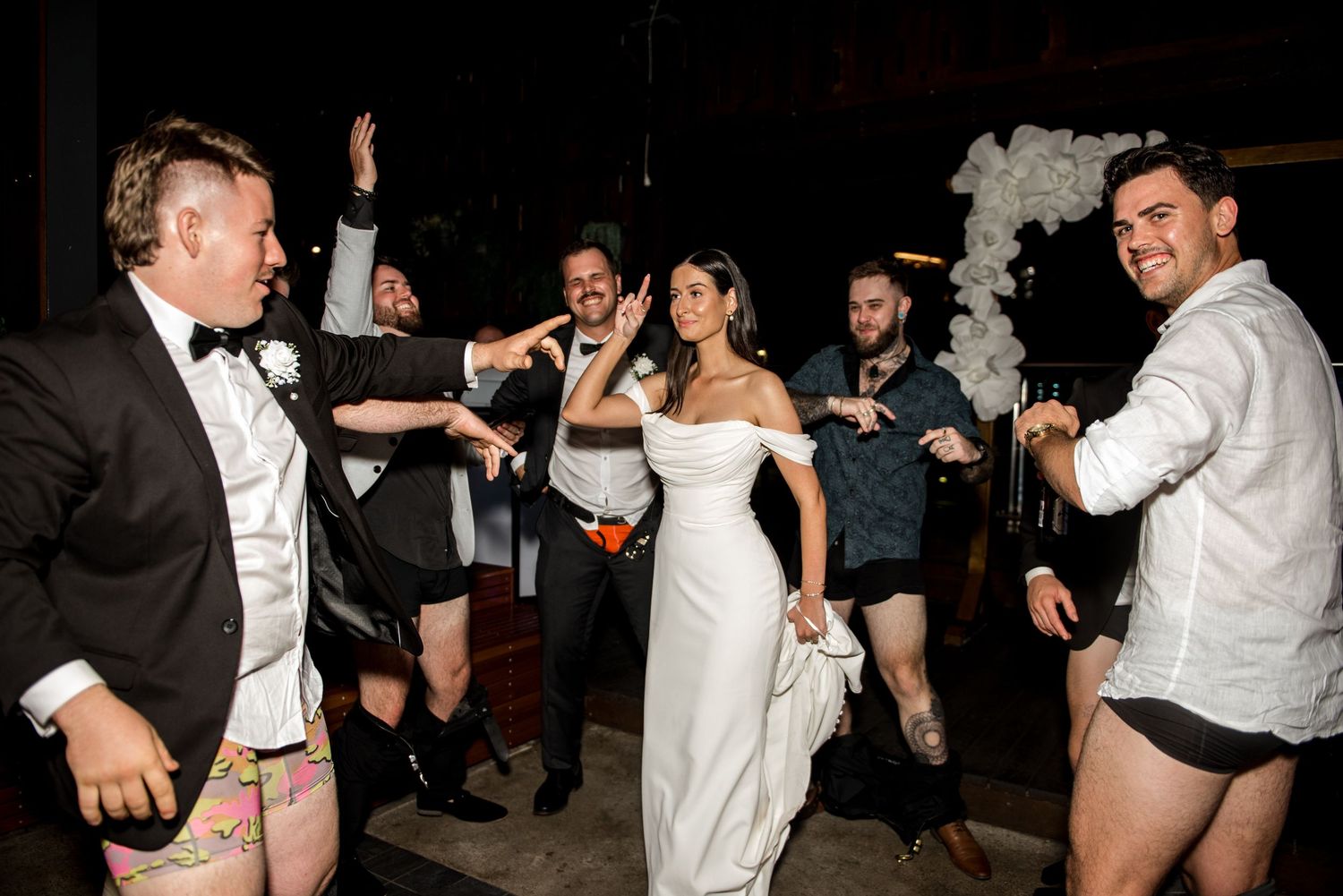 Bride laughs with groomsmen in playful wedding reception dance floor moment with balloon arch backdrop.