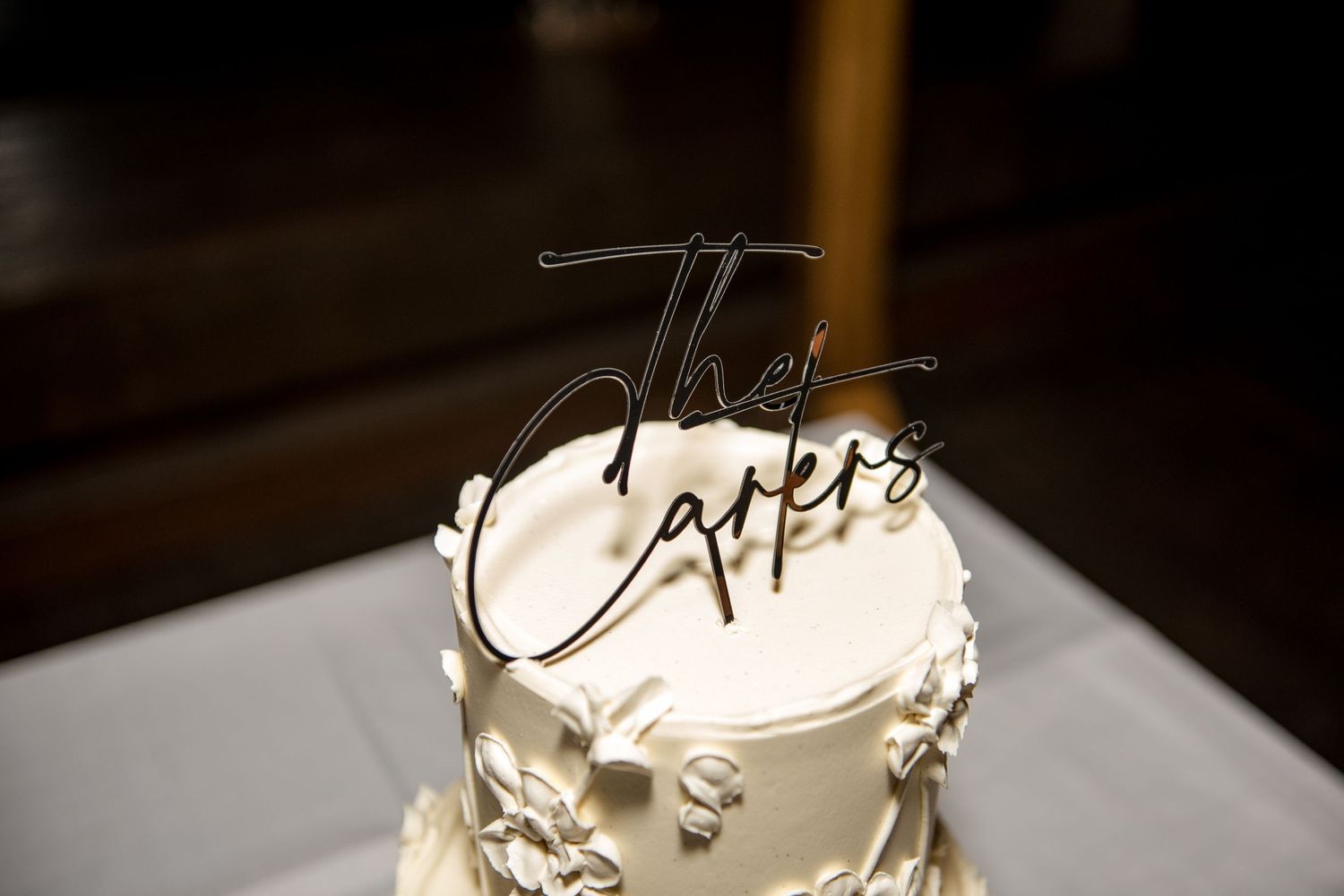 Two-tier white wedding cake topped with a stylish black acrylic script cake topper reading The Carters.