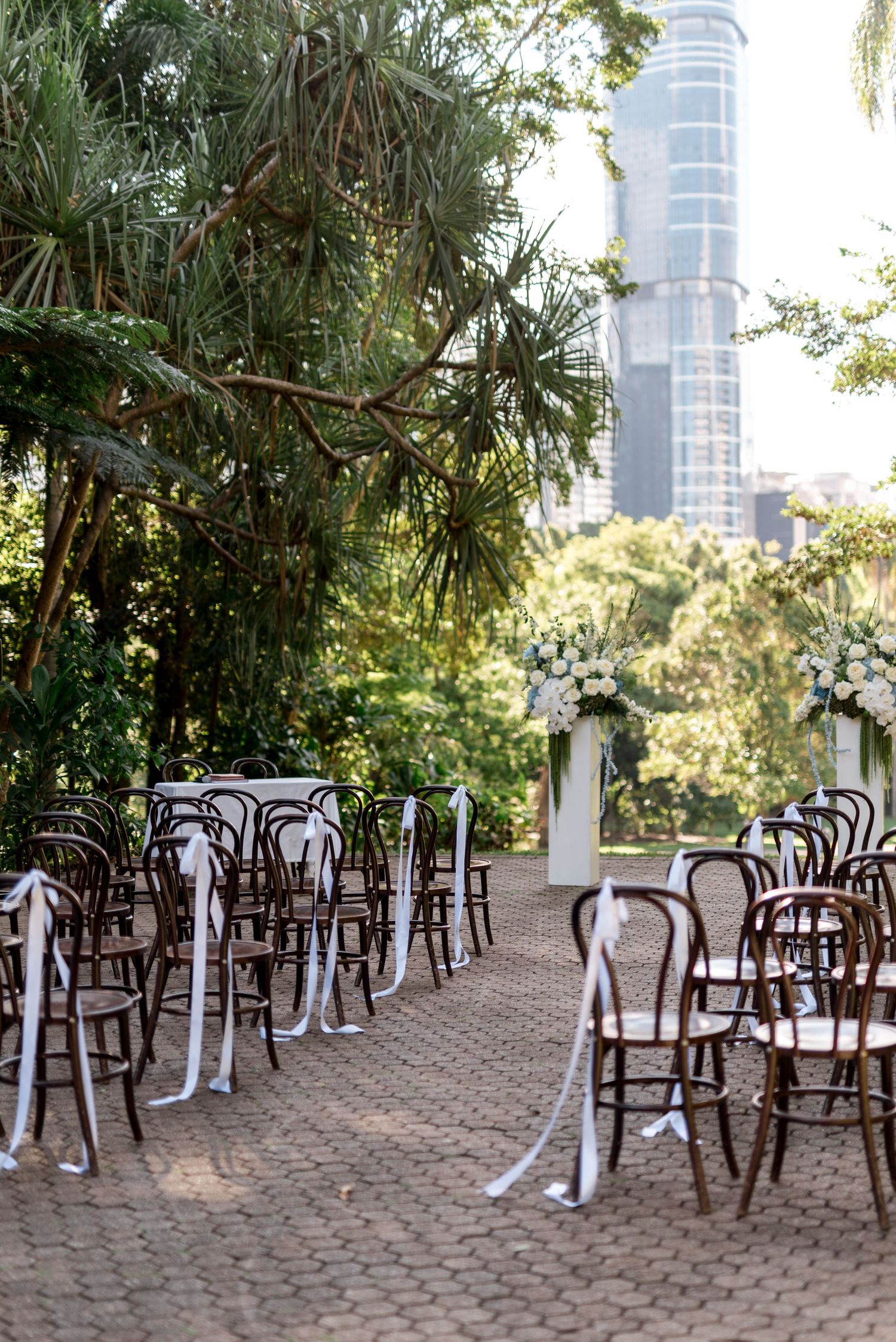 Outdoor wedding ceremony setup with bentwood chairs and white ribbon in a lush garden with city skyscraper backdrop.