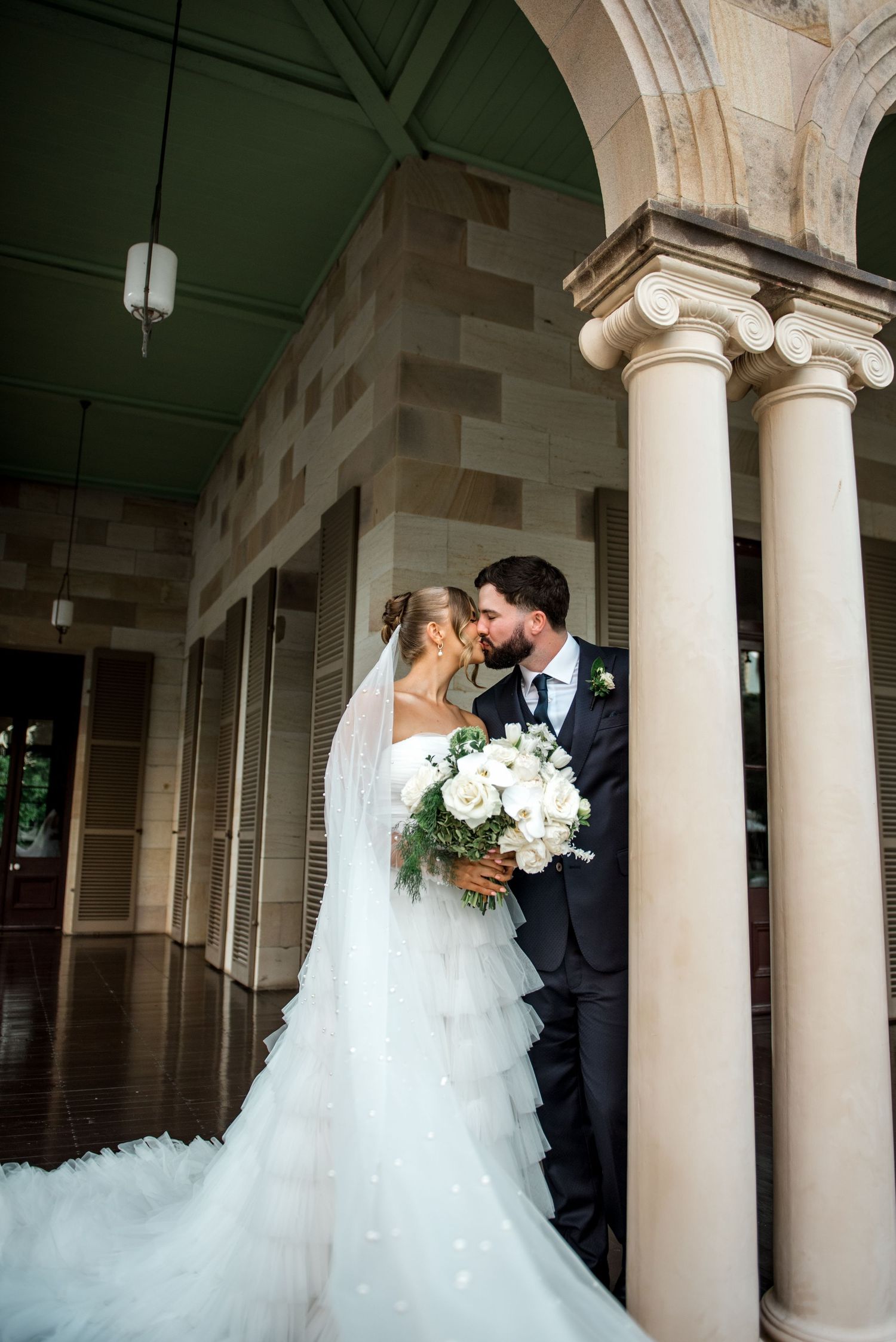 Bride and groom kissing under stone archway with white floral bouquet at wedding venue.