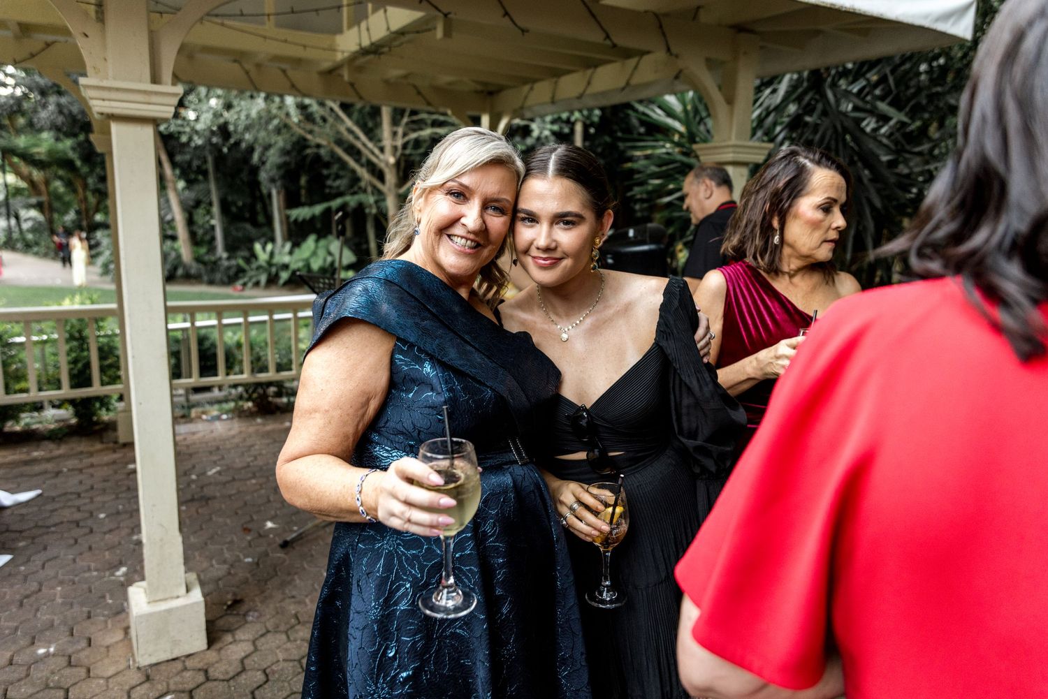 Two women in elegant navy dresses smile and hold champagne glasses at outdoor wedding reception.