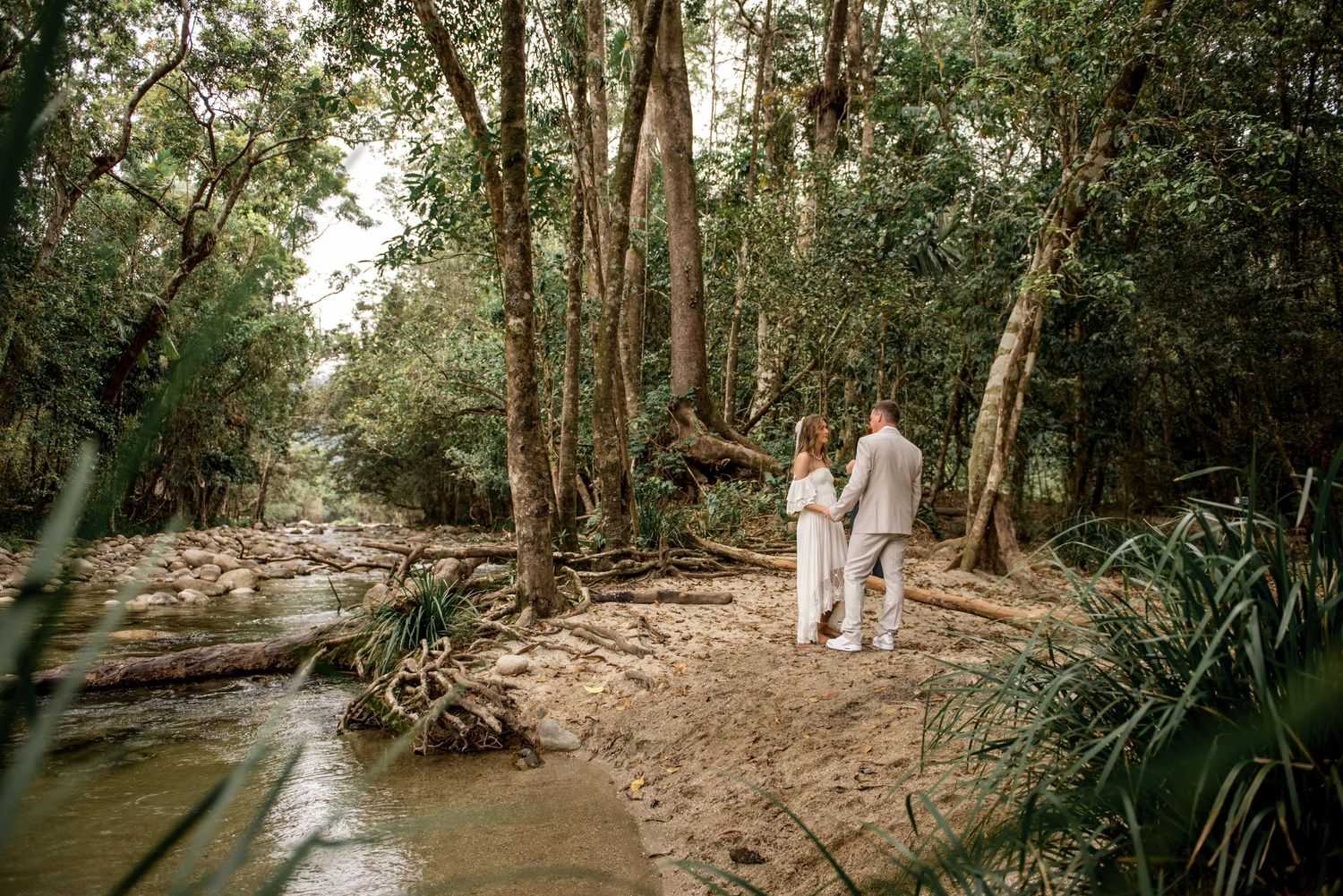 Newlyweds walk hand in hand along a sandy riverbank in a lush rainforest setting.