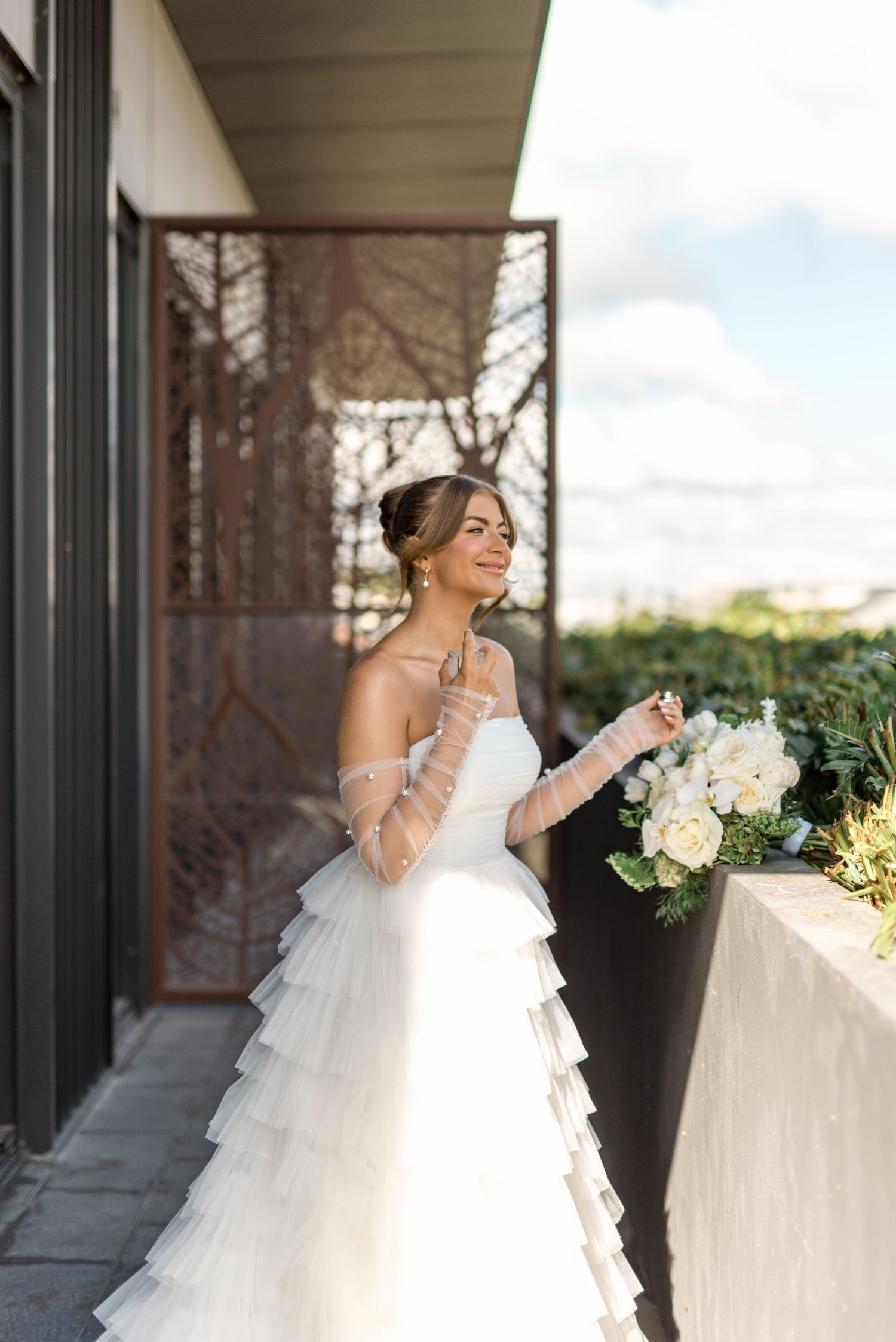 Elegant bride in layered tulle gown with sheer gloves holding white bouquet on sunny outdoor balcony.