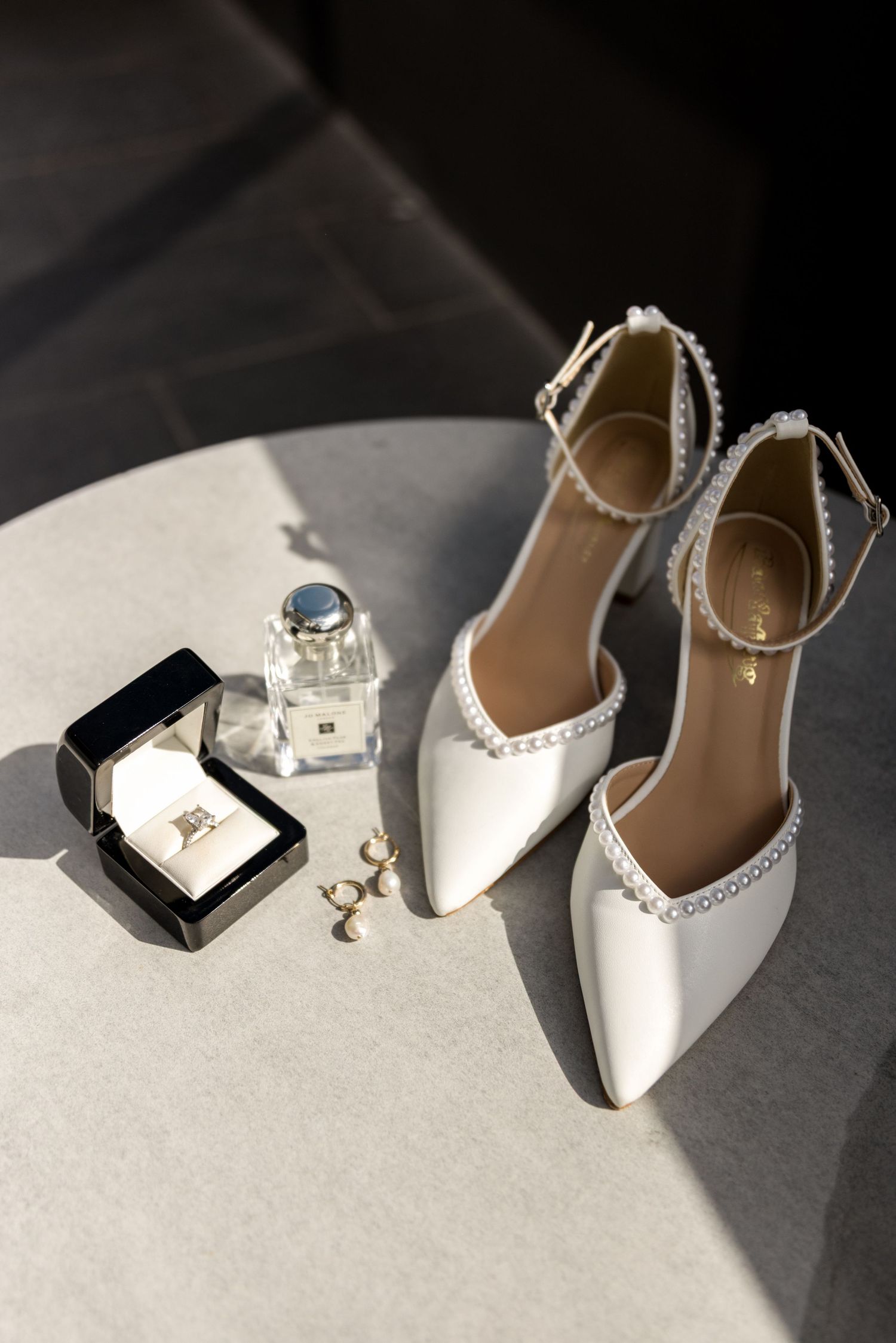 White pearl-trimmed bridal heels displayed beside a diamond ring, earrings, and Jo Malone perfume on a marble table.