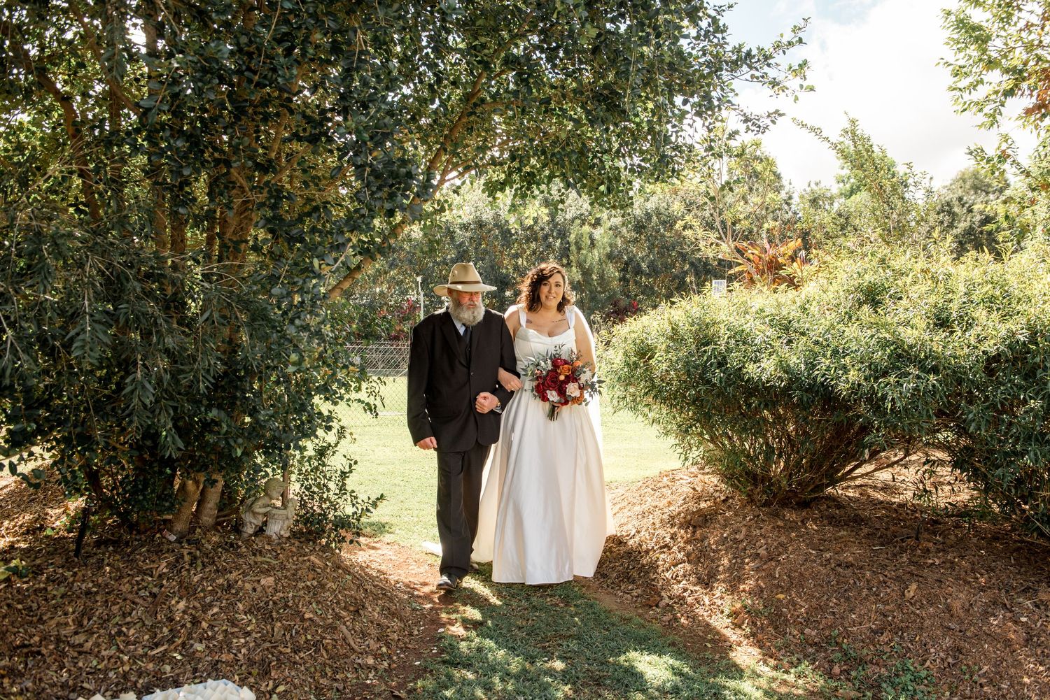 Bride holding red and white bouquet walks with father in hat through sunlit garden pathway.