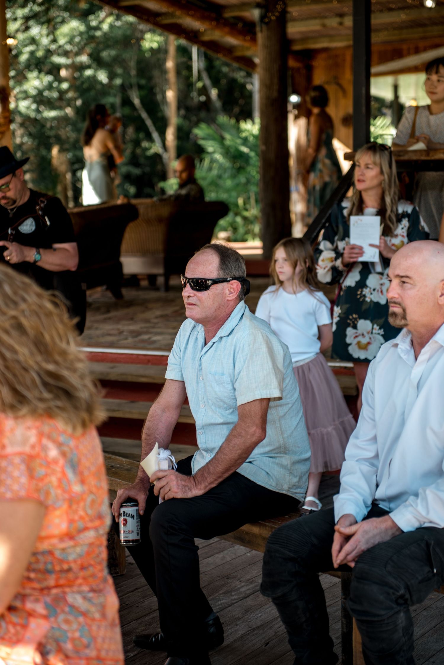 Wedding guests seated outdoors on wooden benches watching ceremony in rustic garden venue.