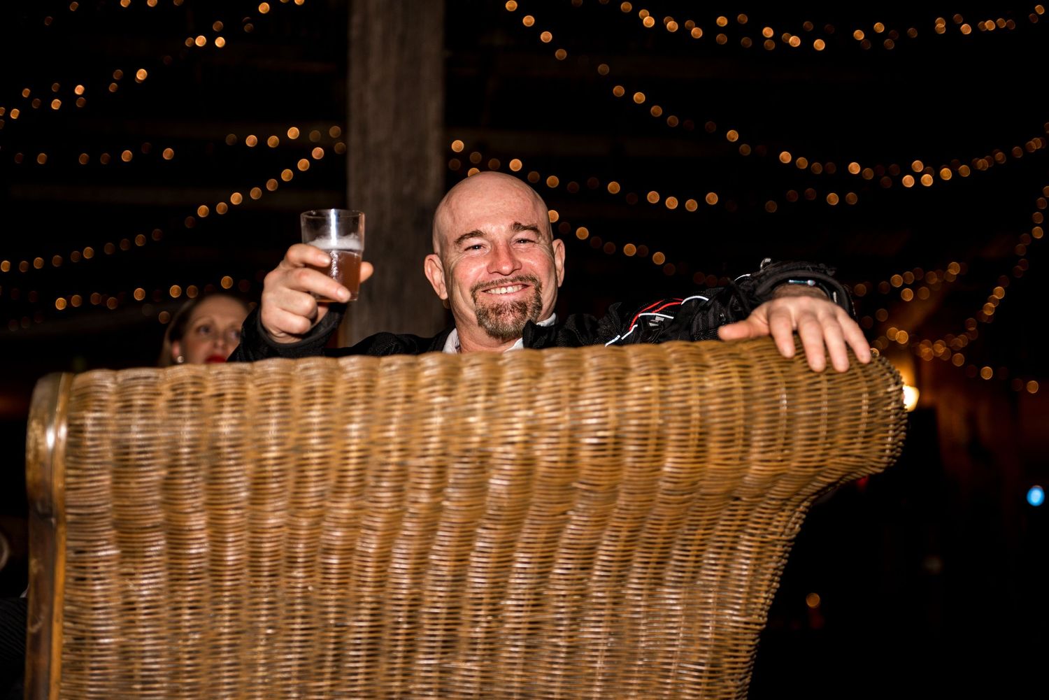 Smiling bald man raises a glass in toast while seated in a wicker chair at wedding.