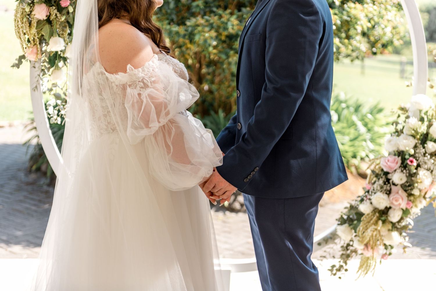Bride and groom hold hands in front of a pink and white floral circular wedding arch.