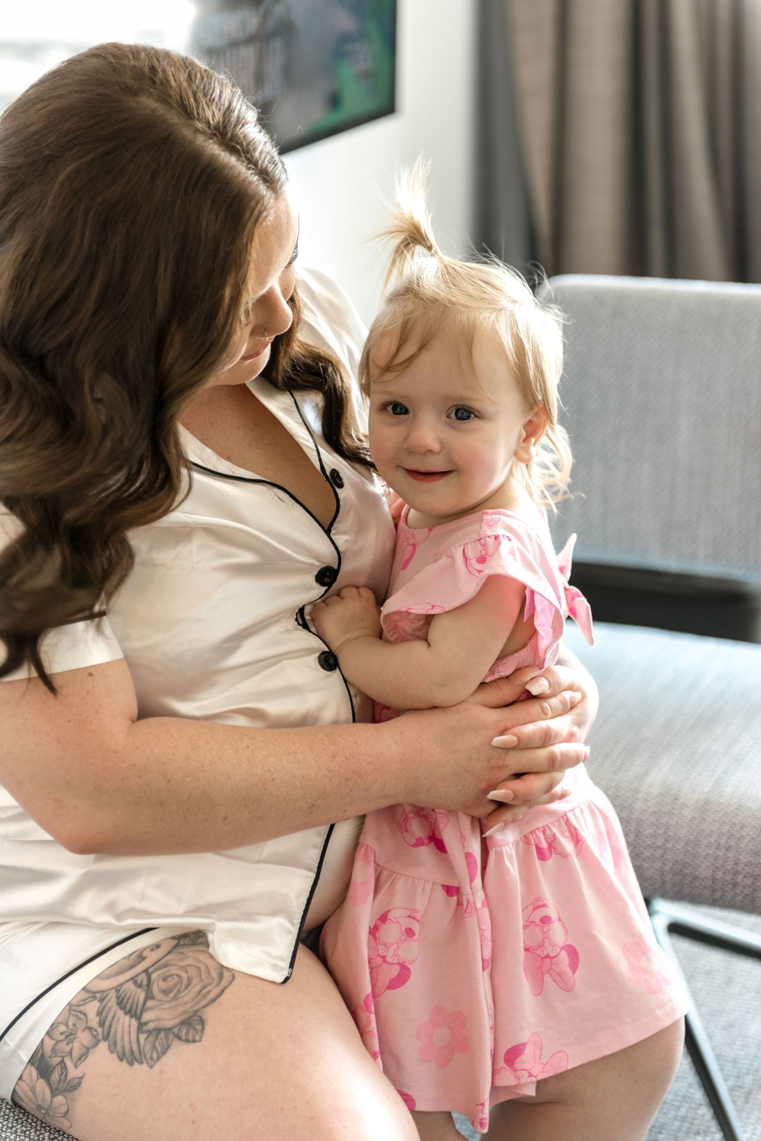Brunette woman in white satin pajamas holding smiling toddler girl in pink floral dress.
