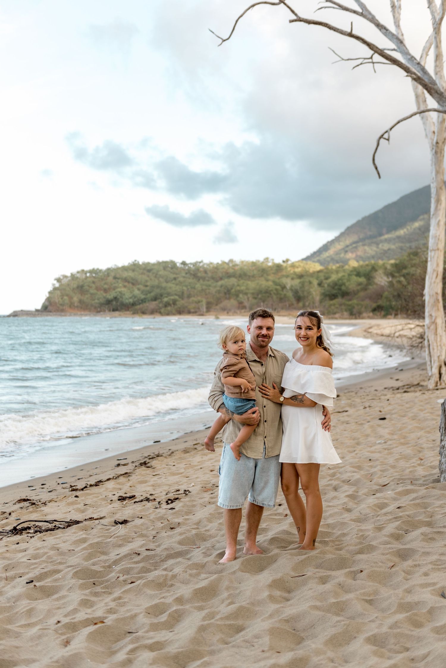 Happy family portrait with toddler on sandy beach beside bare tree with mountain backdrop.