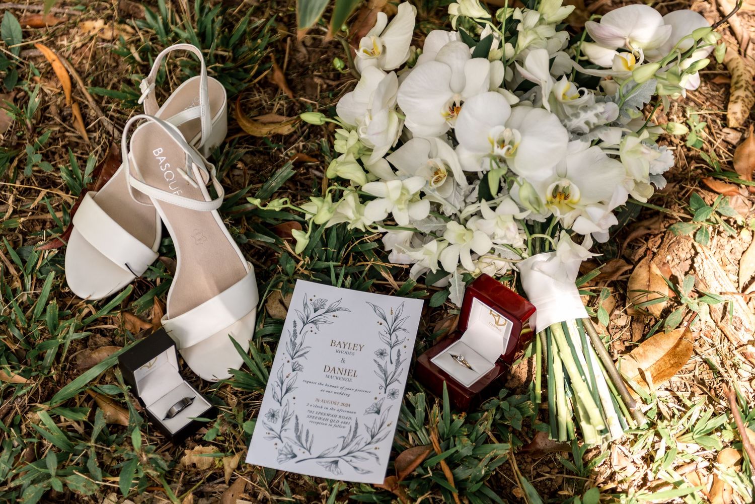 Bridal details flat lay with white sandals, orchid bouquet, wedding invitation, and ring box.