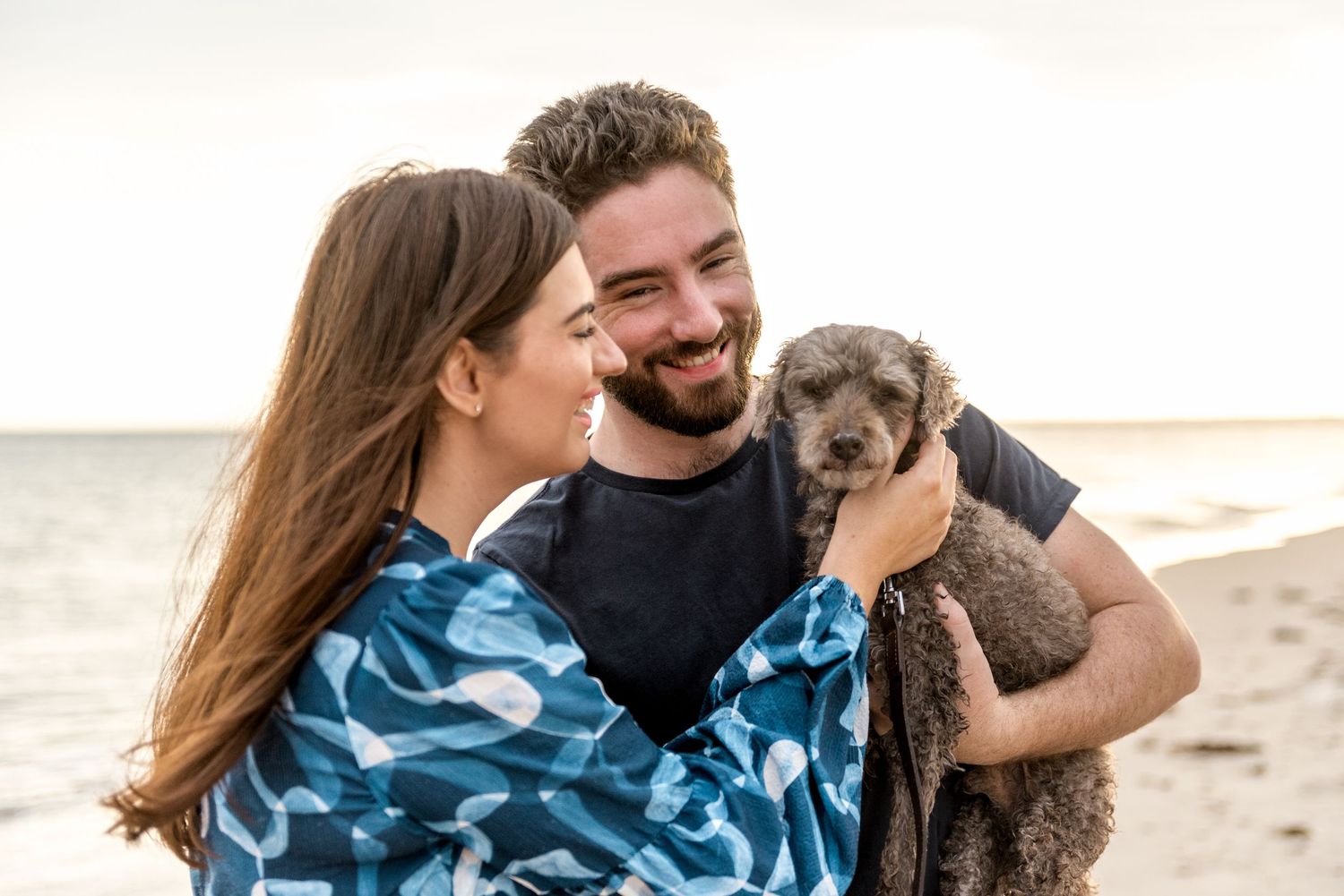 Smiling man holding gray fluffy dog at beach sunset while woman in blue dress looks on.