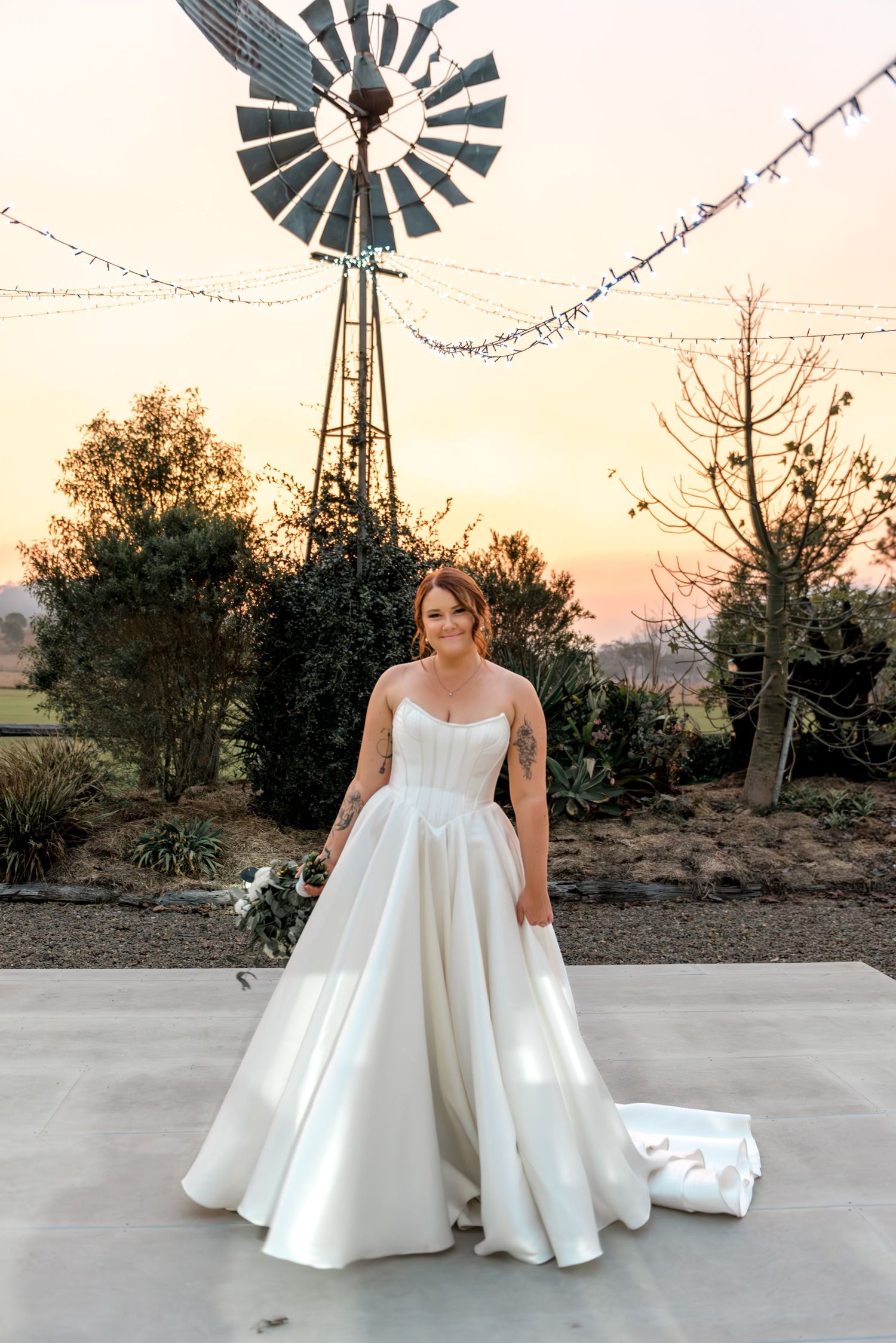 Bride in elegant white satin ballgown poses before a windmill at sunset with string lights overhead.