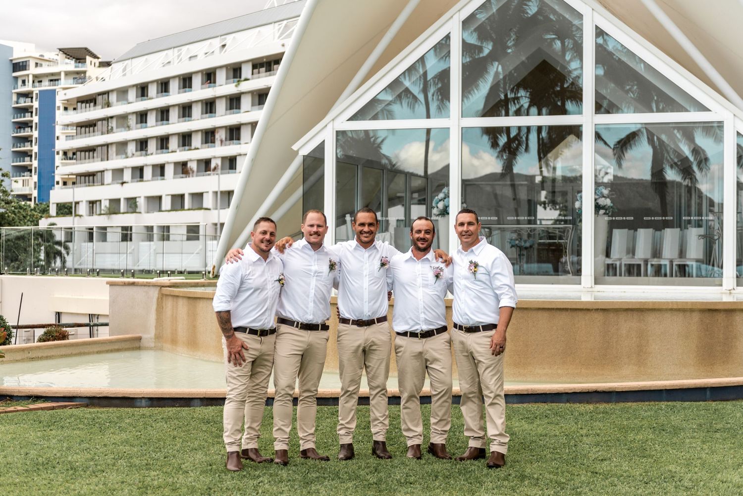 Five groomsmen in matching white shirts and khaki pants posing outside a glass chapel building.