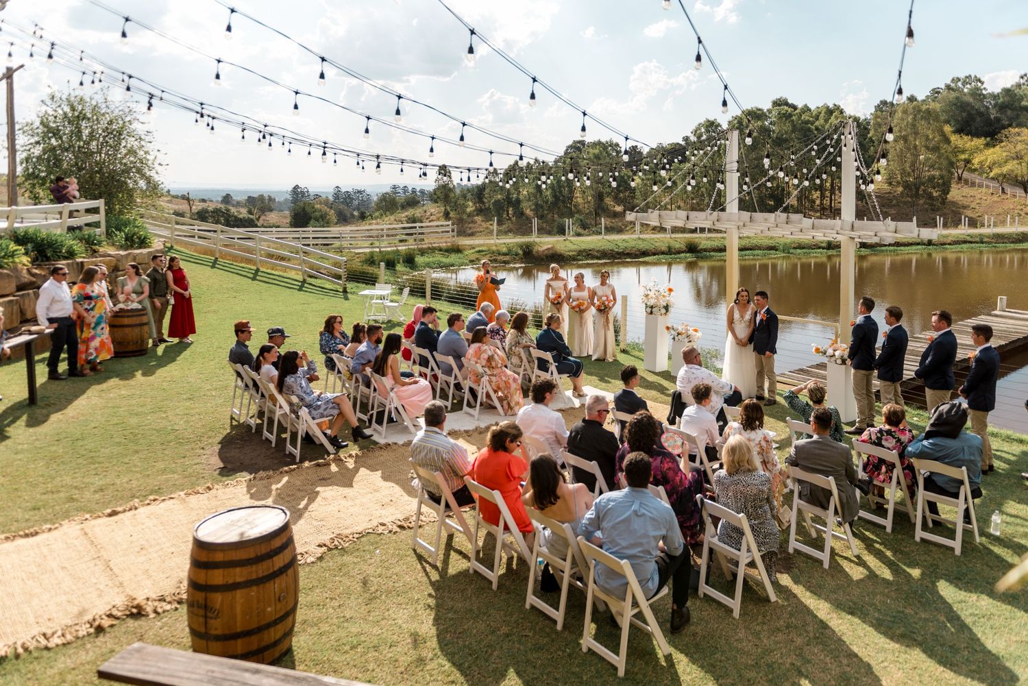 Outdoor wedding ceremony with guests seated on white chairs near a pond, decorated with string lights overhead.