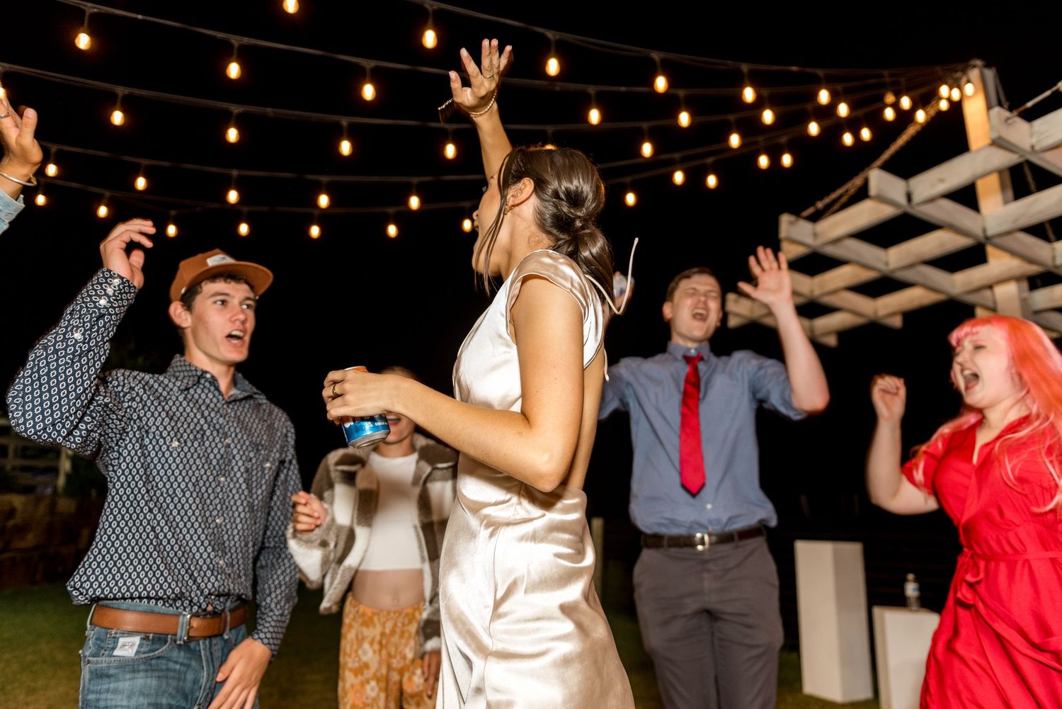 Wedding guests dancing energetically outdoors under string lights at night reception.