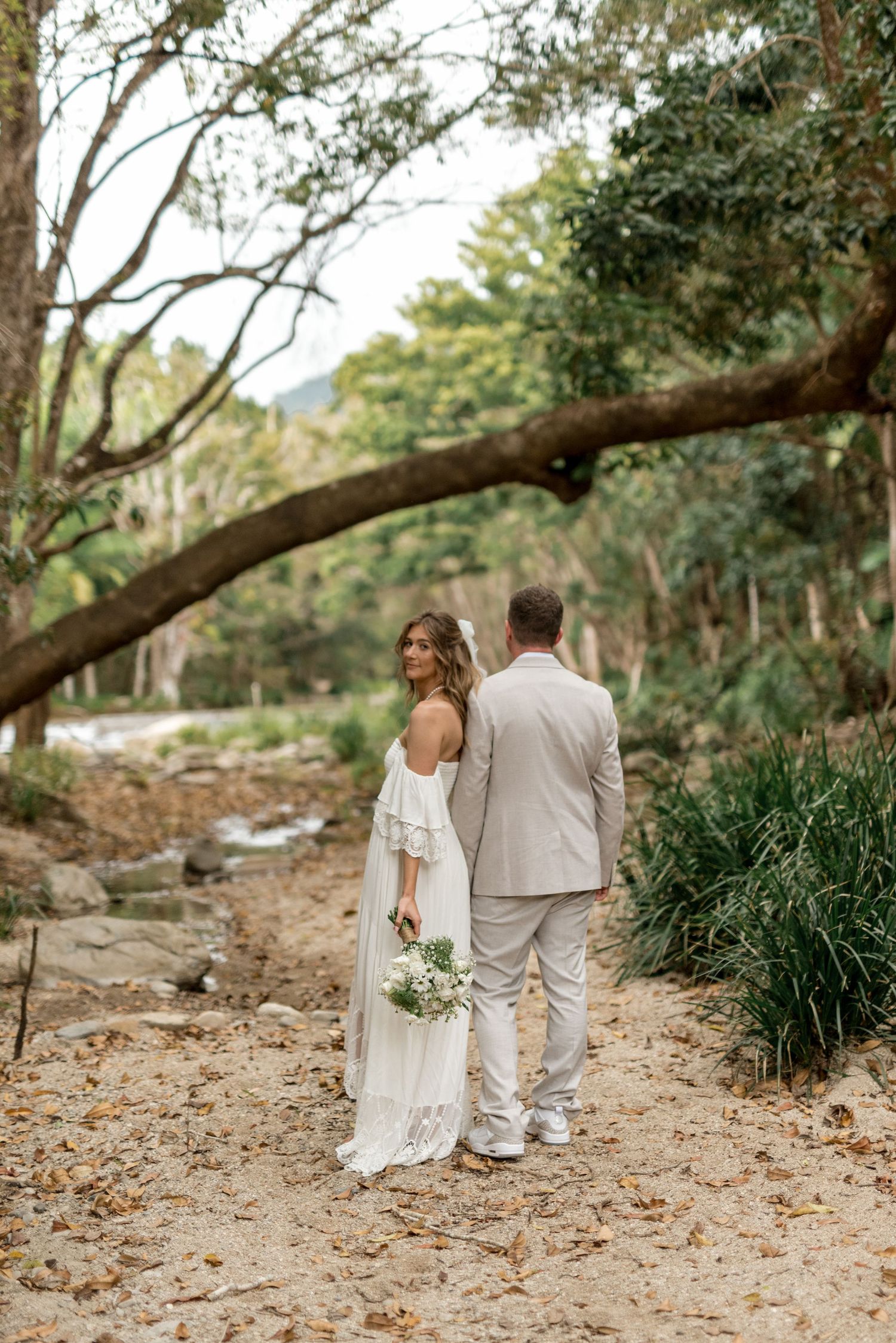 Bride holding bouquet and groom walk away under a sweeping tree branch in a forest setting.
