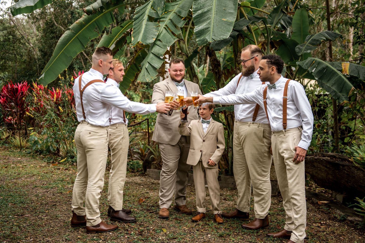 Groomsmen and ring bearer toasting with beer glasses in tropical garden before wedding.