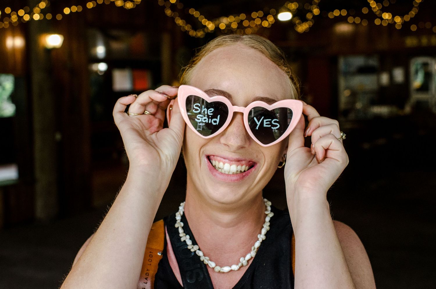 Smiling wedding guest holding pink heart-shaped sunglasses reading She Said Yes.