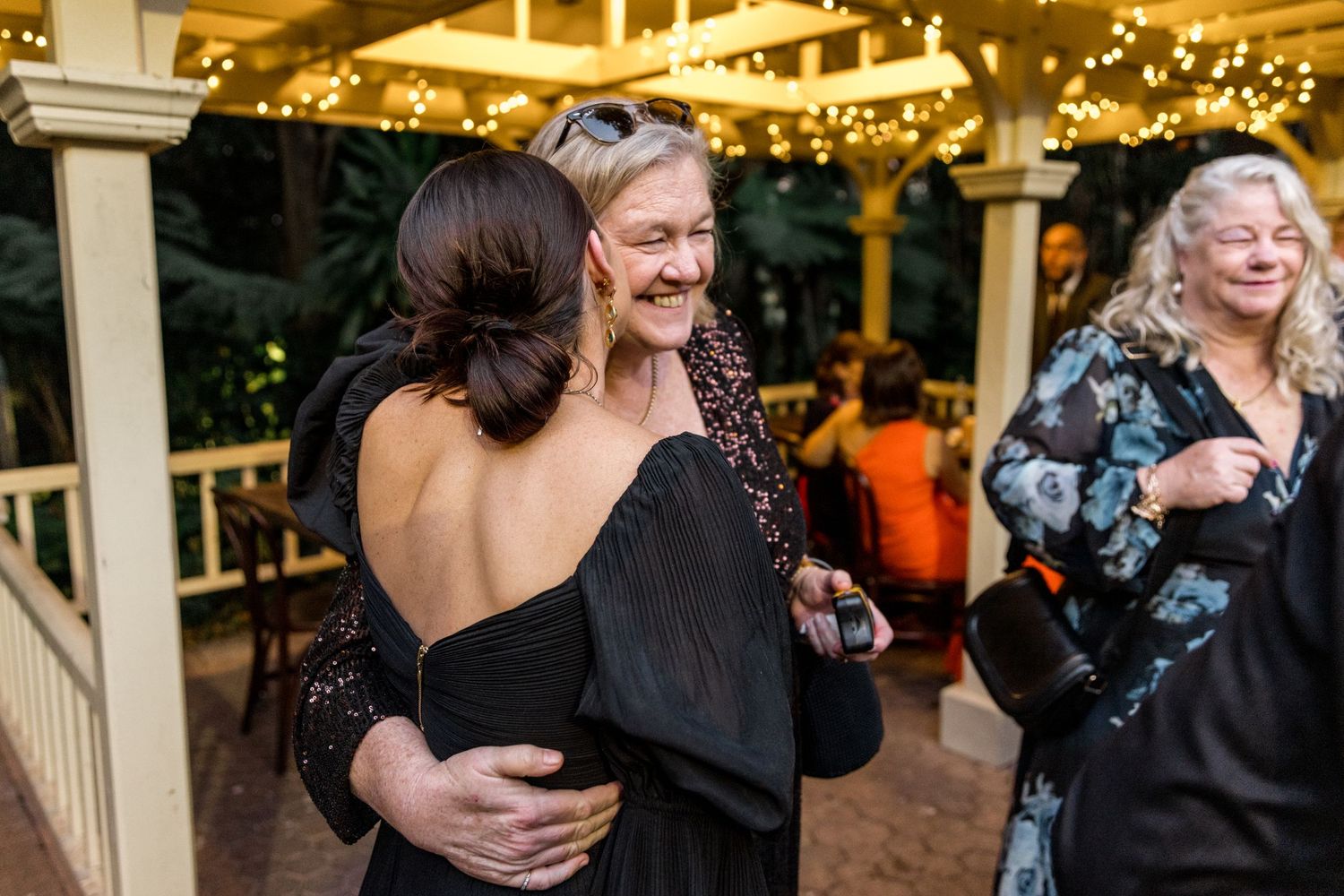 Two women share a warm embrace at an outdoor wedding reception with string lights.