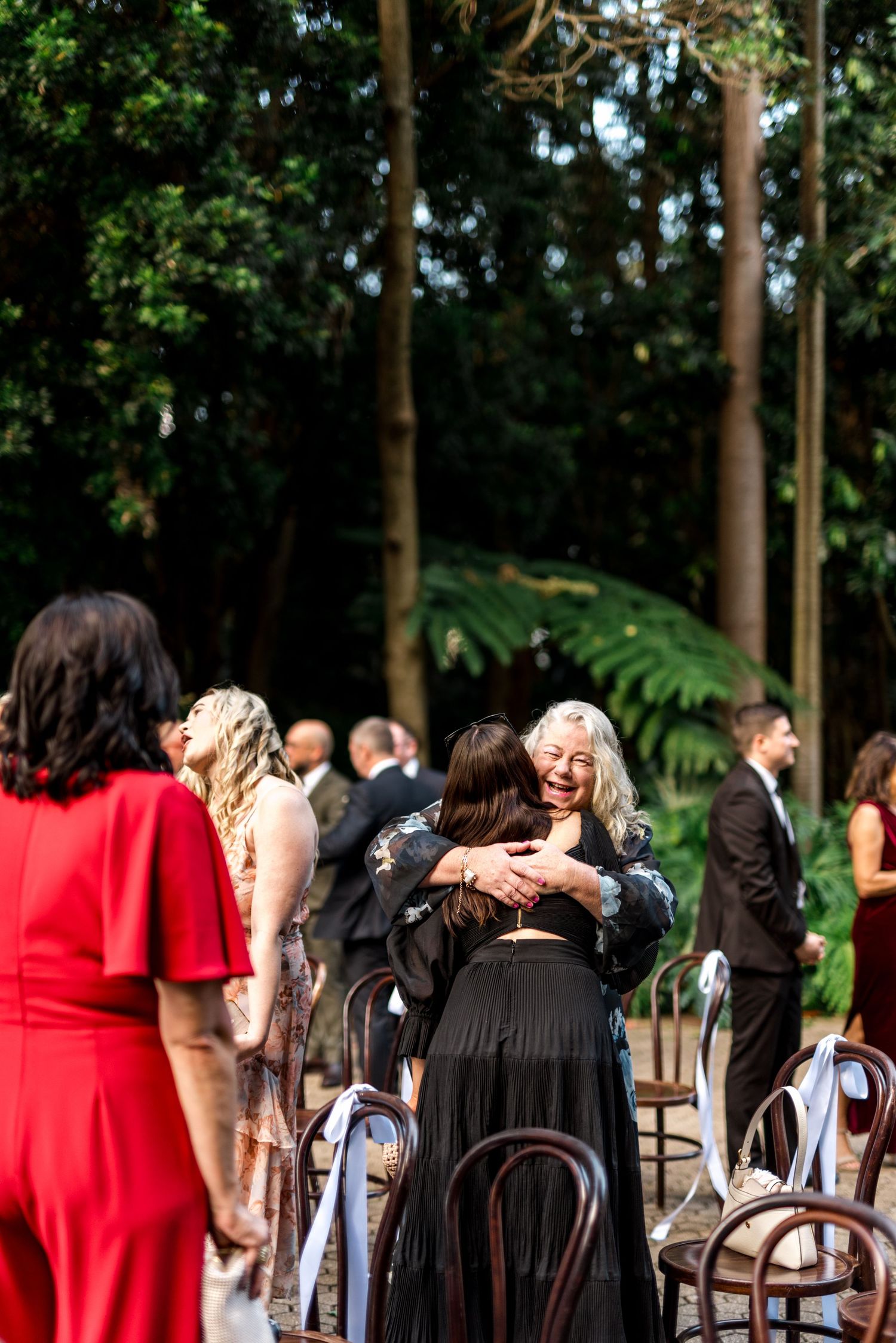 Guests embrace warmly after an outdoor garden wedding ceremony among tall trees.