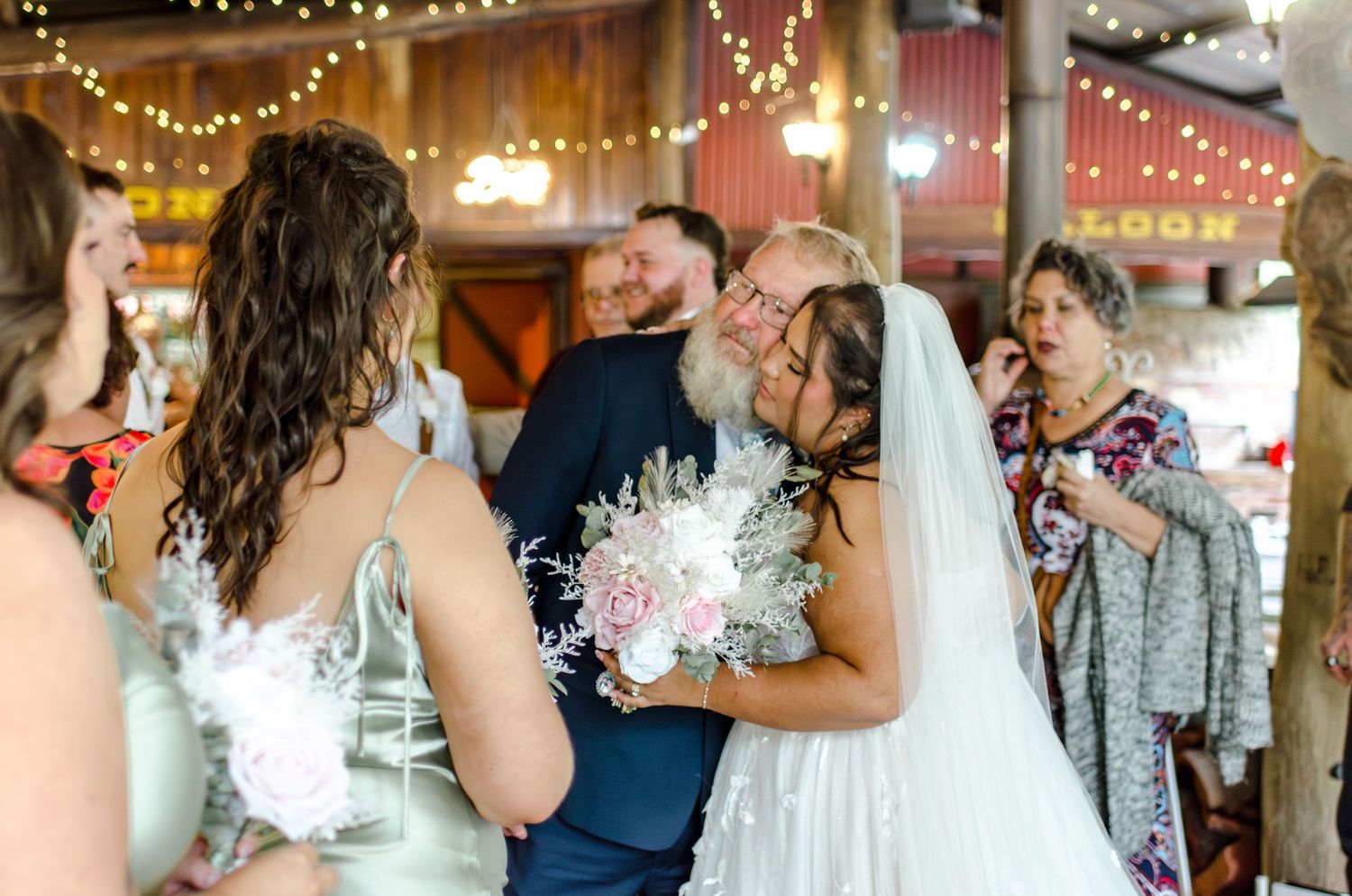 Bride hugs a guest while holding a white floral bouquet inside a rustic barn wedding venue.