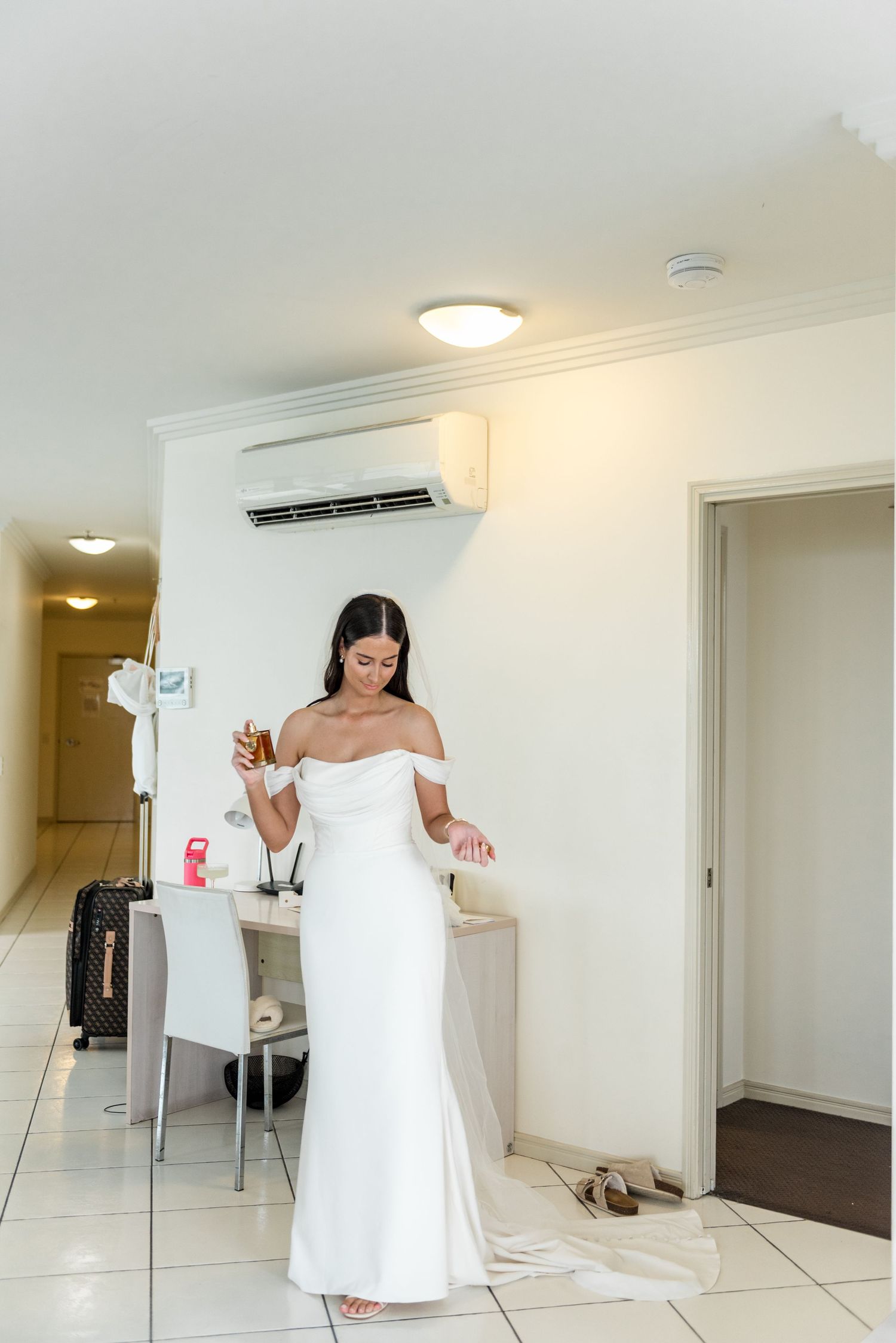 Bride in off-shoulder white gown holding a drink while getting ready in a hotel room.