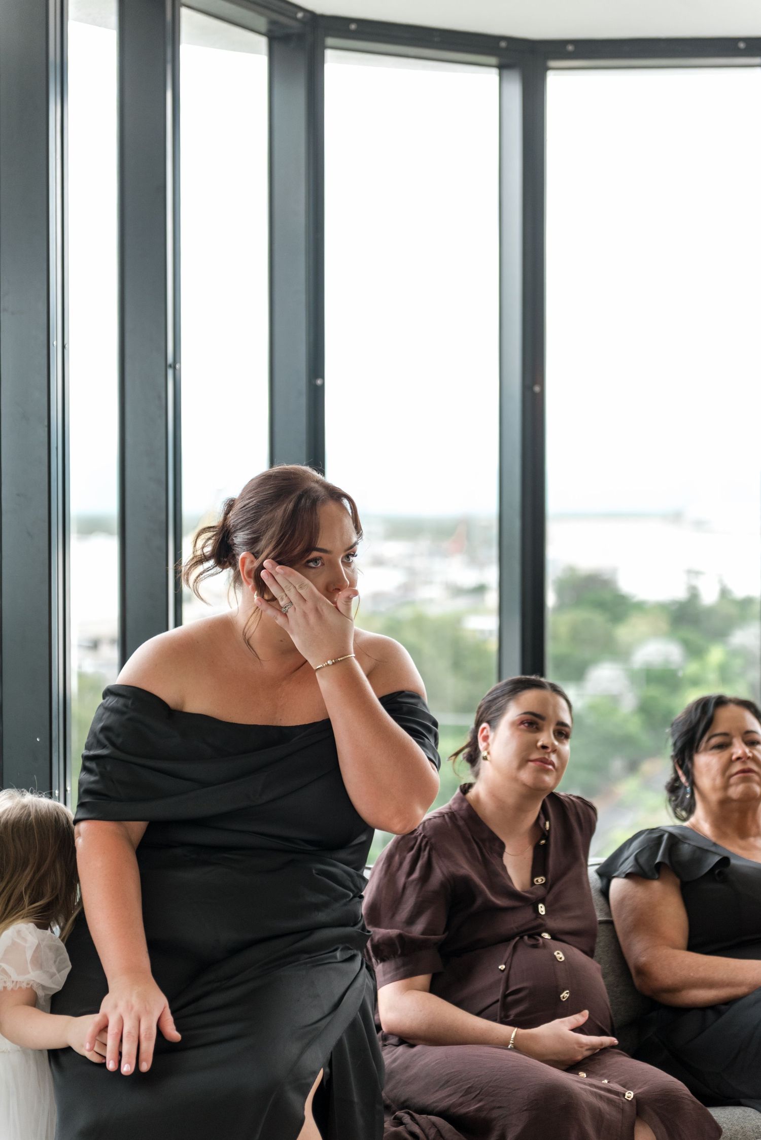 An emotional bridesmaid in a black off-shoulder dress wipes a tear while seated guests react to the bride's reveal.
