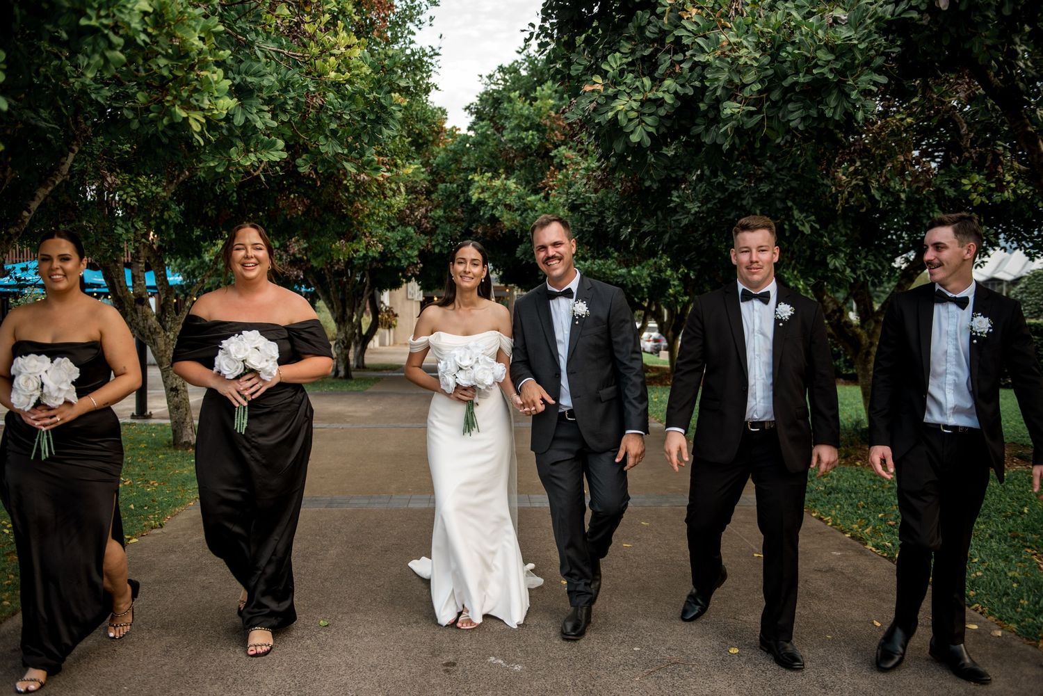 Smiling wedding party walking together along a tree-lined park pathway.