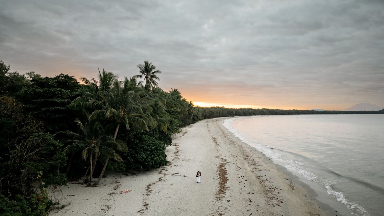 Aerial view of tropical beach at sunset with palm trees and cloudy sky.