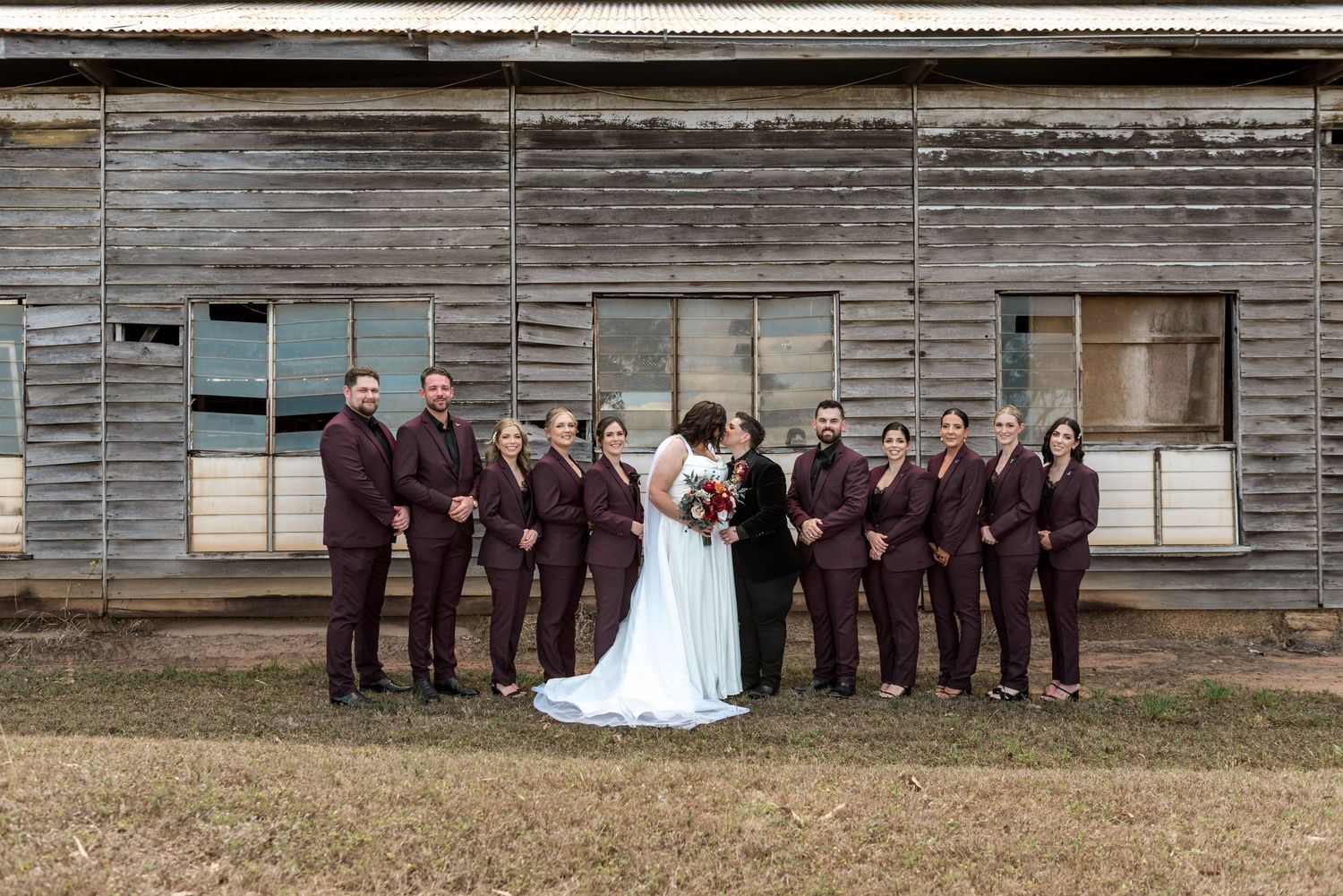 Wedding party in coordinating burgundy suits poses with bride and groom before a weathered wooden barn exterior.