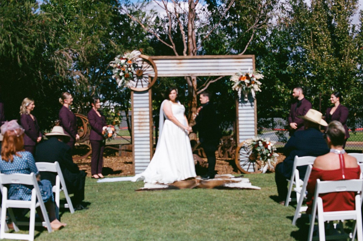 Bride and groom exchange vows at an outdoor ceremony with a rustic arch and guests seated on white chairs.