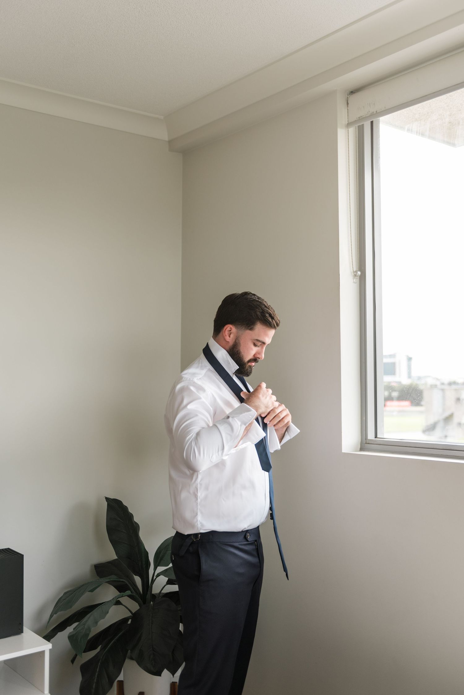 Groom adjusting his navy tie while getting dressed in a white shirt near a bright window.