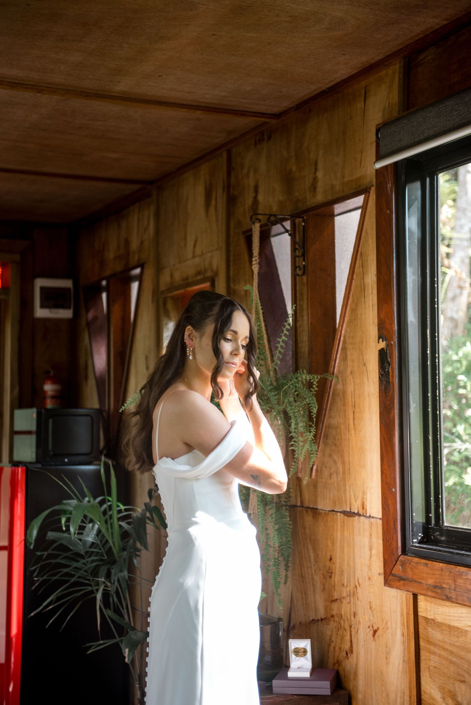 Bride in elegant off-shoulder white gown posing in sunlit rustic wooden interior.
